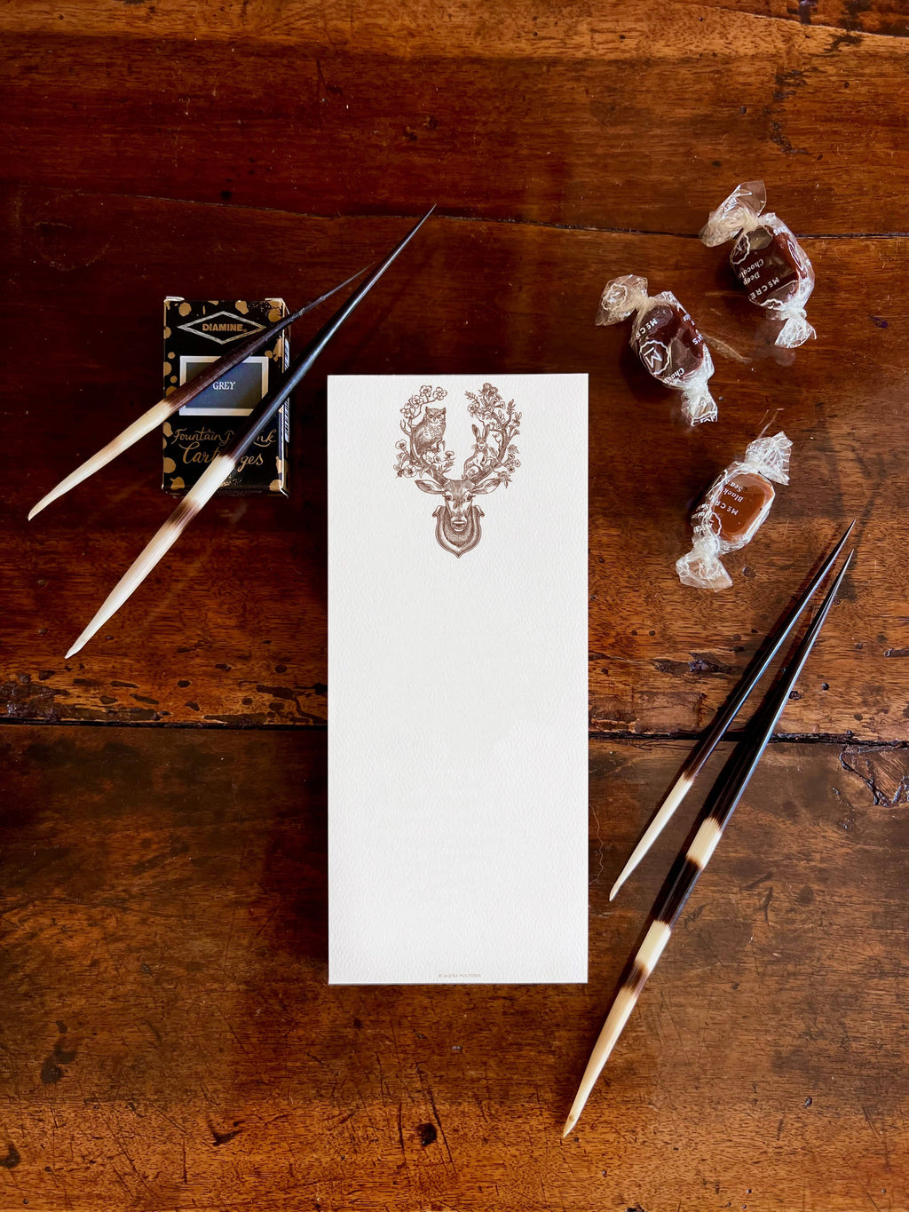 A white letterhead card with a brown printed crest sits on a rustic dark wood table, surrounded by matching black-and-cream writing tools and wrapped chocolates.