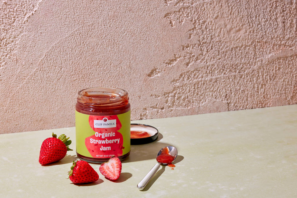 Jar of Clif Family Napa Valley organic strawberry jam with fresh strawberries and a rustic label on a wooden surface.