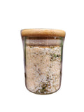 A clear glass jar filled with a light beige and green herbal or spice blend, topped with a natural bamboo lid, displayed on a rustic wooden surface with green leaves in the background.