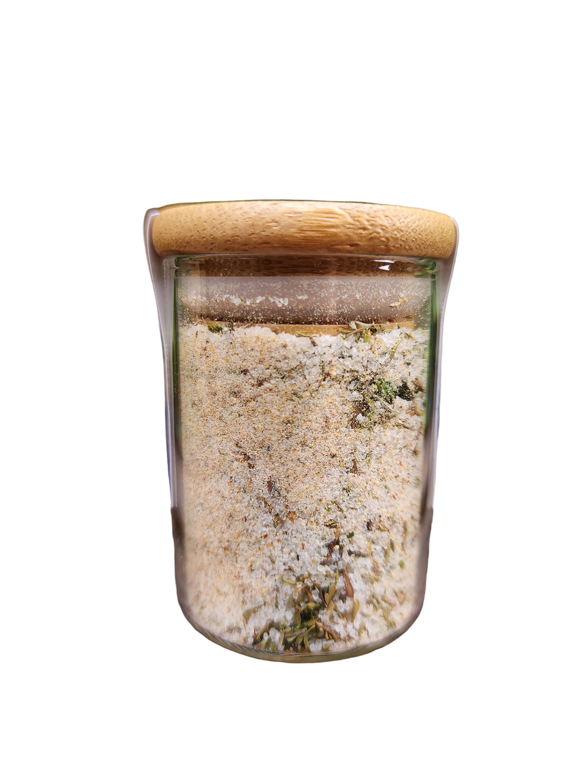 A clear glass jar filled with a light beige and green herbal or spice blend, topped with a natural bamboo lid, displayed on a rustic wooden surface with green leaves in the background.