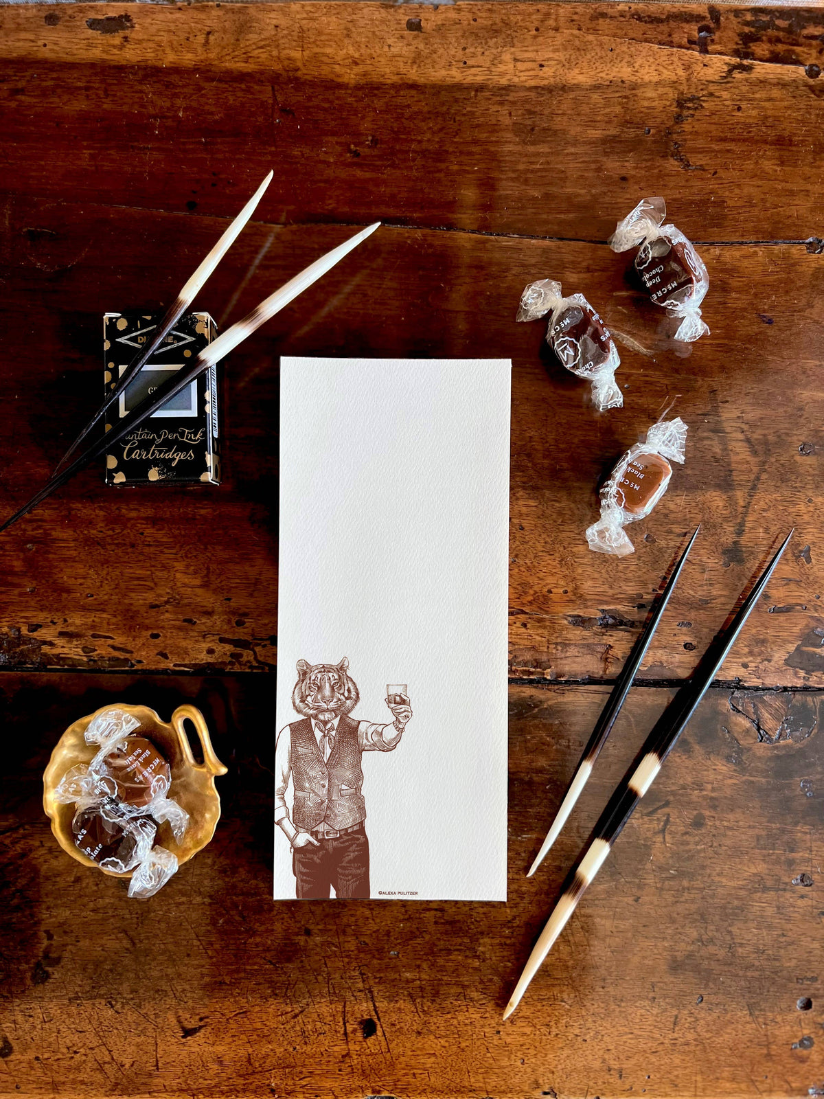 Wooden surface with a notepad featuring a tiger holding a glass, surrounded by decorative items.