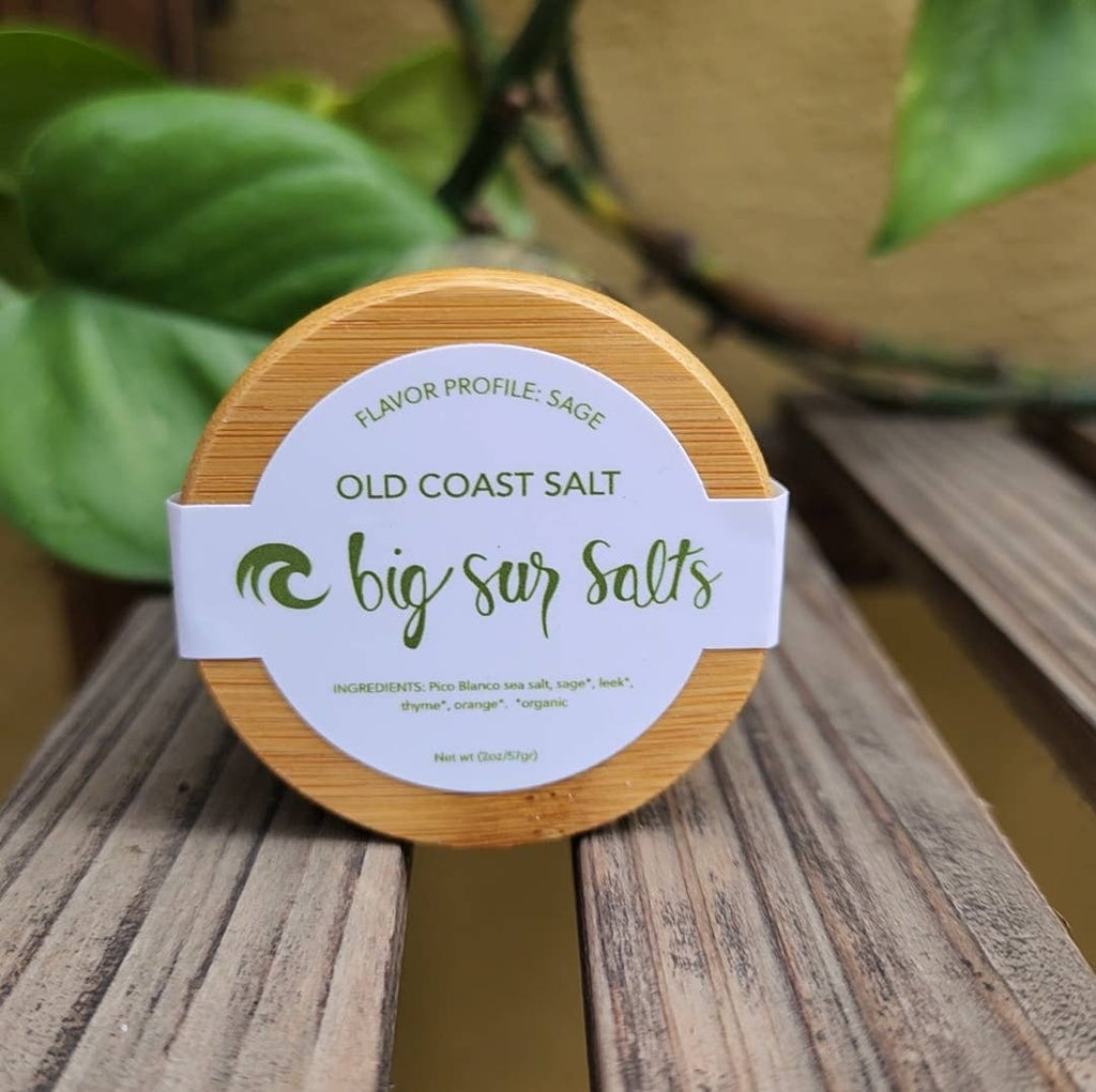A round wooden container of Big Sur Salts Old Coast Salt with a white paper band label, resting on a weathered wooden surface with green foliage in the background.