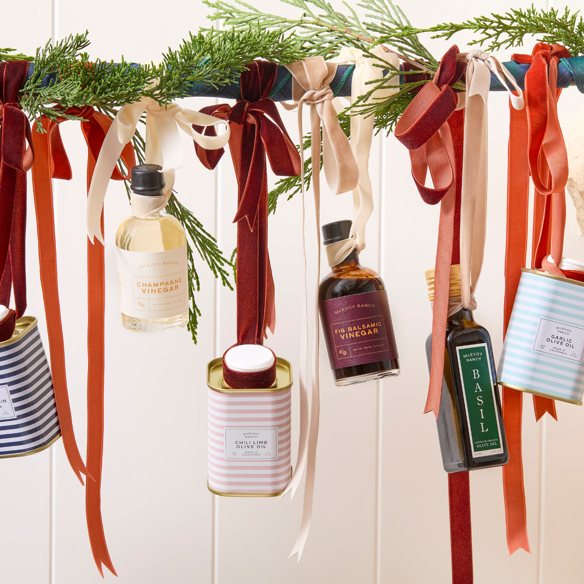 A festive holiday hanging display with green pine branches draped across a white paneled wall, adorned with colorful ribbons and several decorative bottles and tins labeled with holiday flavors like champagne vinegar, tragedy vinegar, basil, and nut oil.