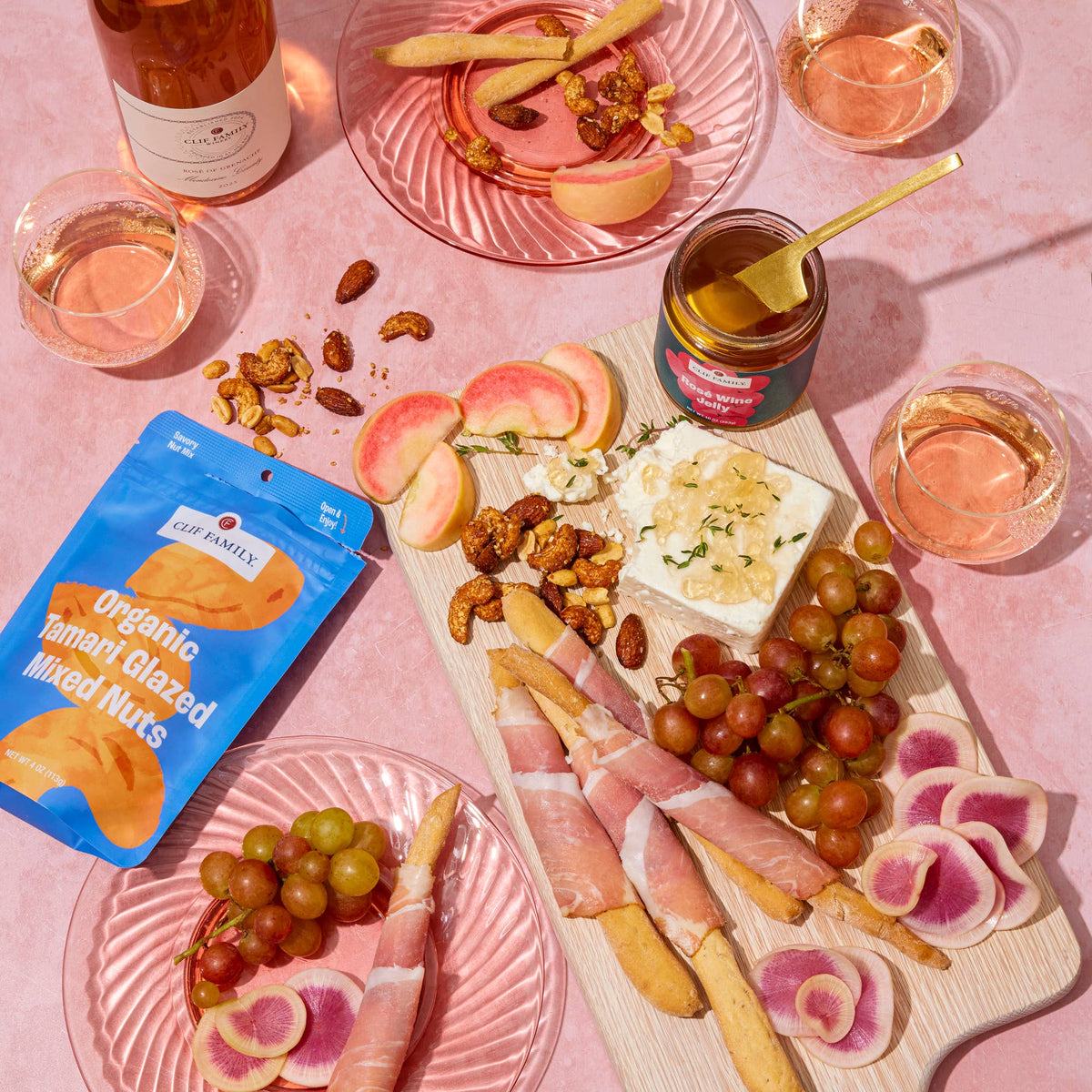 A styled charcuterie board scene on a pink surface with a wooden board holding cheese, grapes, nuts, prosciutto, crackers, and a jar of mixed nuts. A blue bag of Clif Family Organic Turmeric Glazed Mixed Nuts is visible, along with pink plates, glasses of rosé wine, and a bottle in the background.