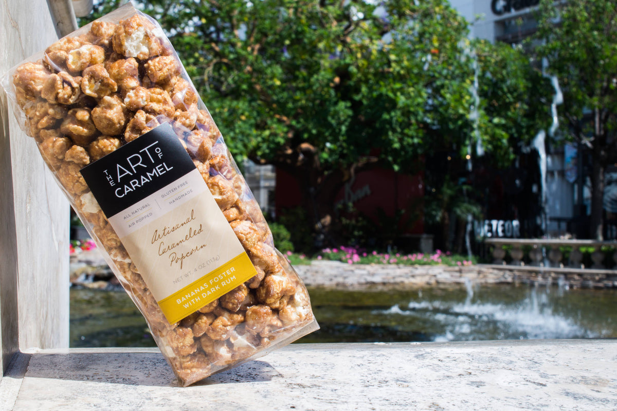A clear bag of Art of Caramel caramelized apricot popcorn leaning against a stone ledge outdoors, with a blurred pond and trees in the background.