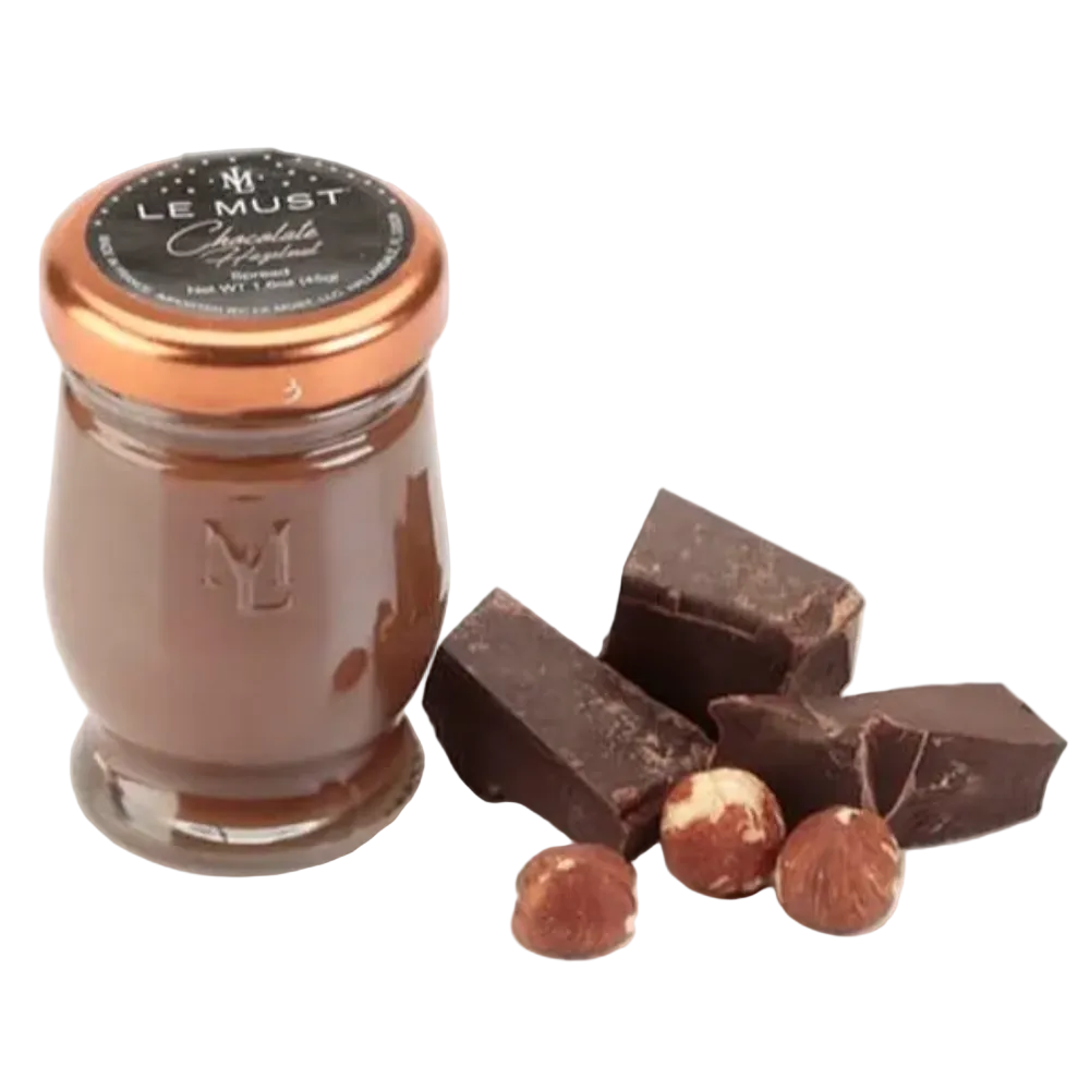 A small glass jar of Le Must Chocolate Hazelnut spread with a copper-colored lid and black label, shown next to pieces of dark chocolate and hazelnuts on a white background.