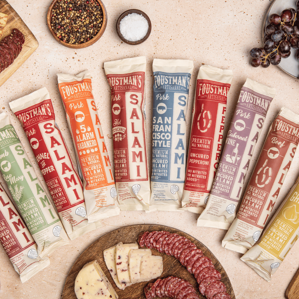 Assorted Foustman's Salami packages on a table with salami slices and cheese.