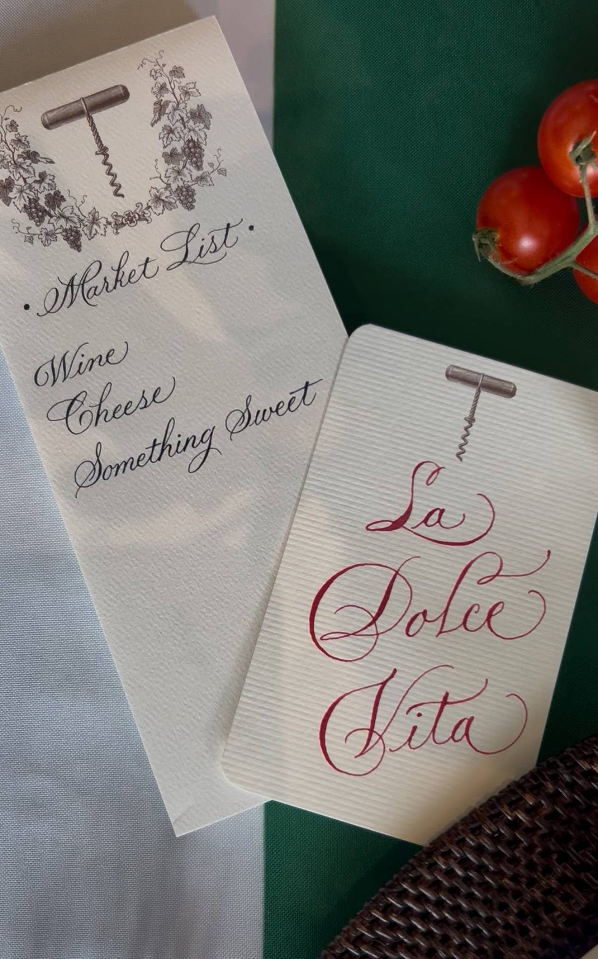 Two cream-colored paper place cards with corkscrew illustrations, one printed with black cursive text reading 'Market List' and the other with red cursive text reading 'La Dolce Vita', styled on a dark green surface with tomatoes and a woven basket edge.