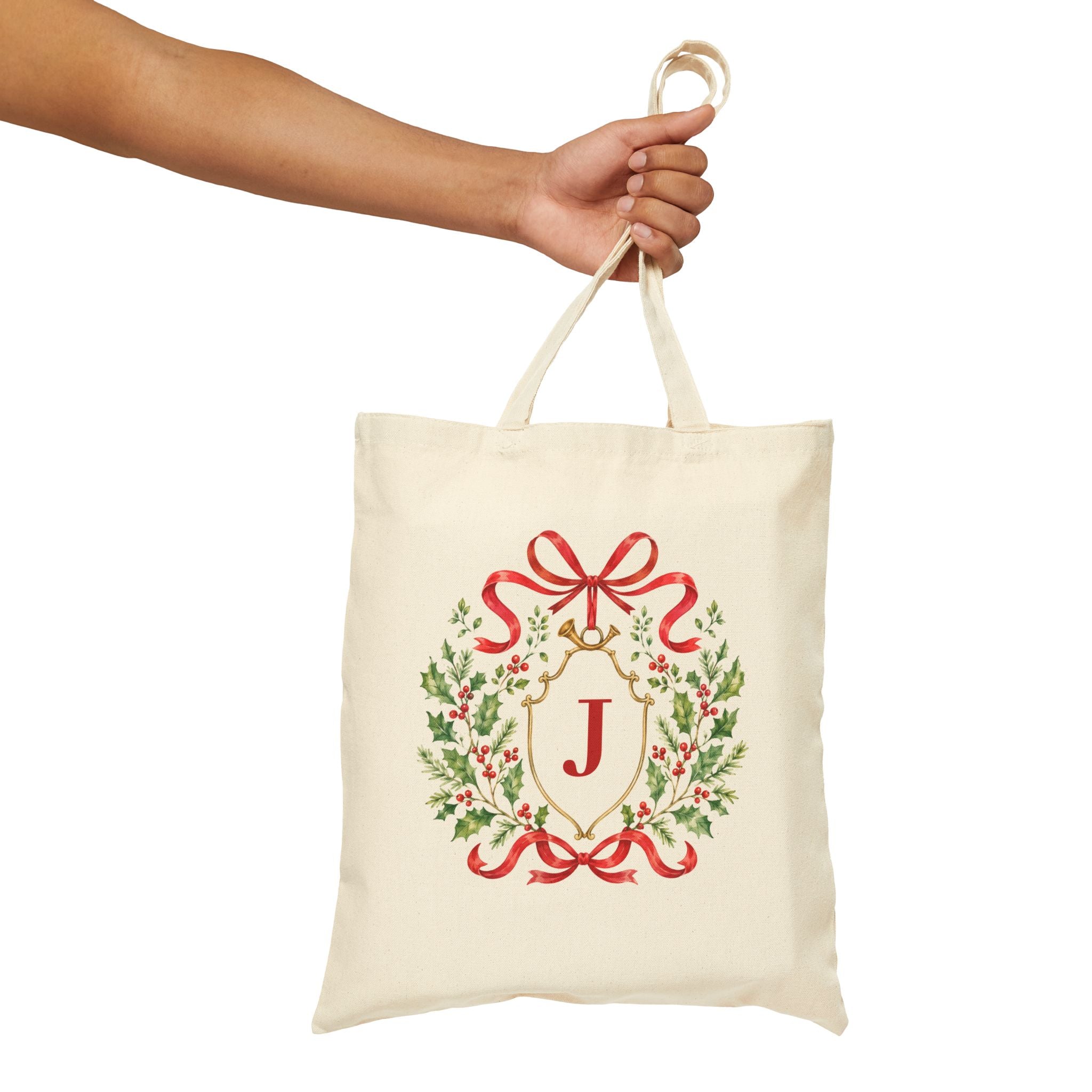 An off-white canvas tote bag with a red and green Christmas wreath design featuring a gold monogram letter 'J' and a red ribbon bow, held by a person's hand against a white background.