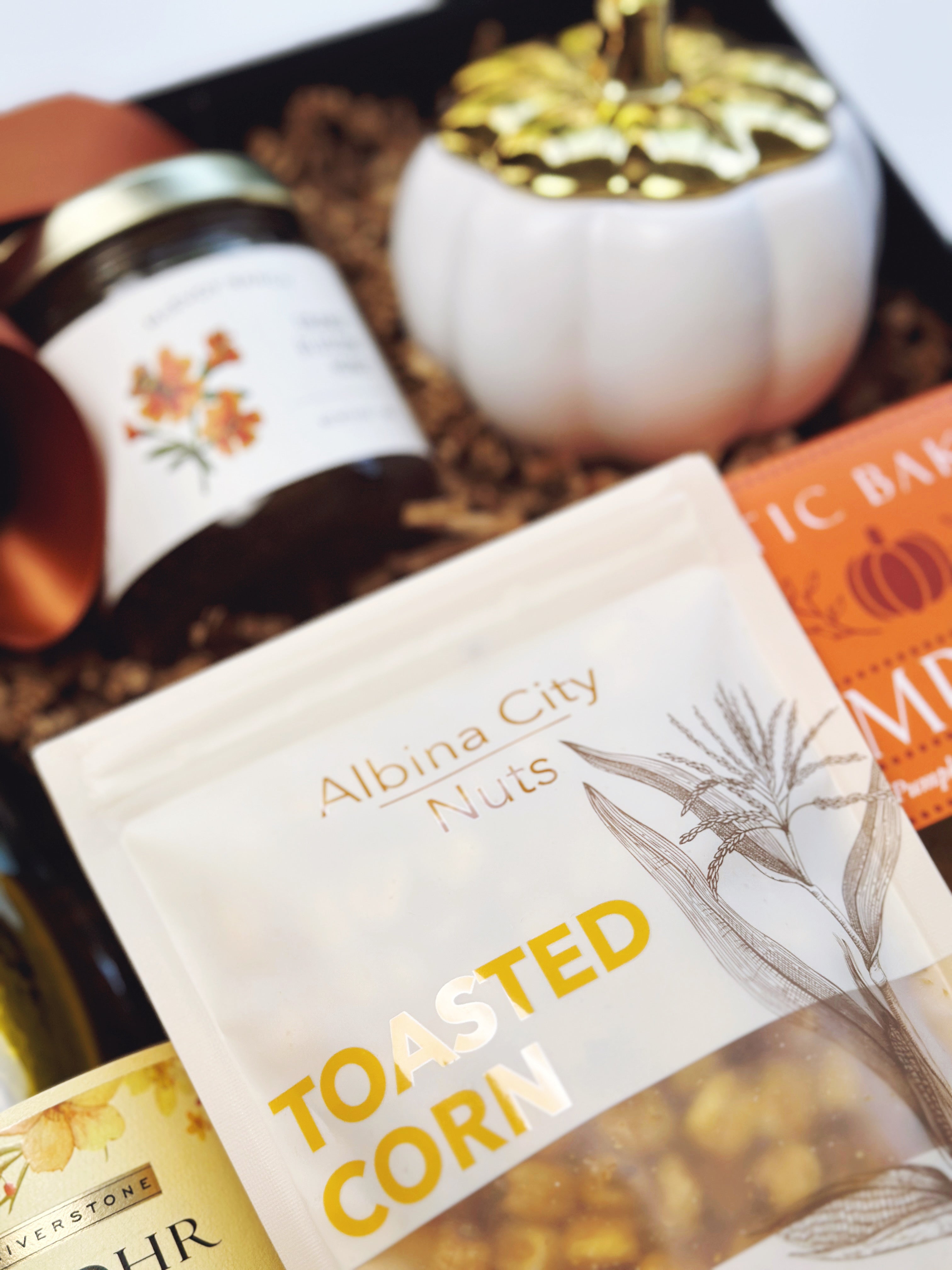 A styled gift basket or tray containing Albina City Nuts Toasted Corn in a clear bag with yellow lettering, a jar of honey,  a white decorative pumpkin candle, arranged in a black gift box with rust ribbon.