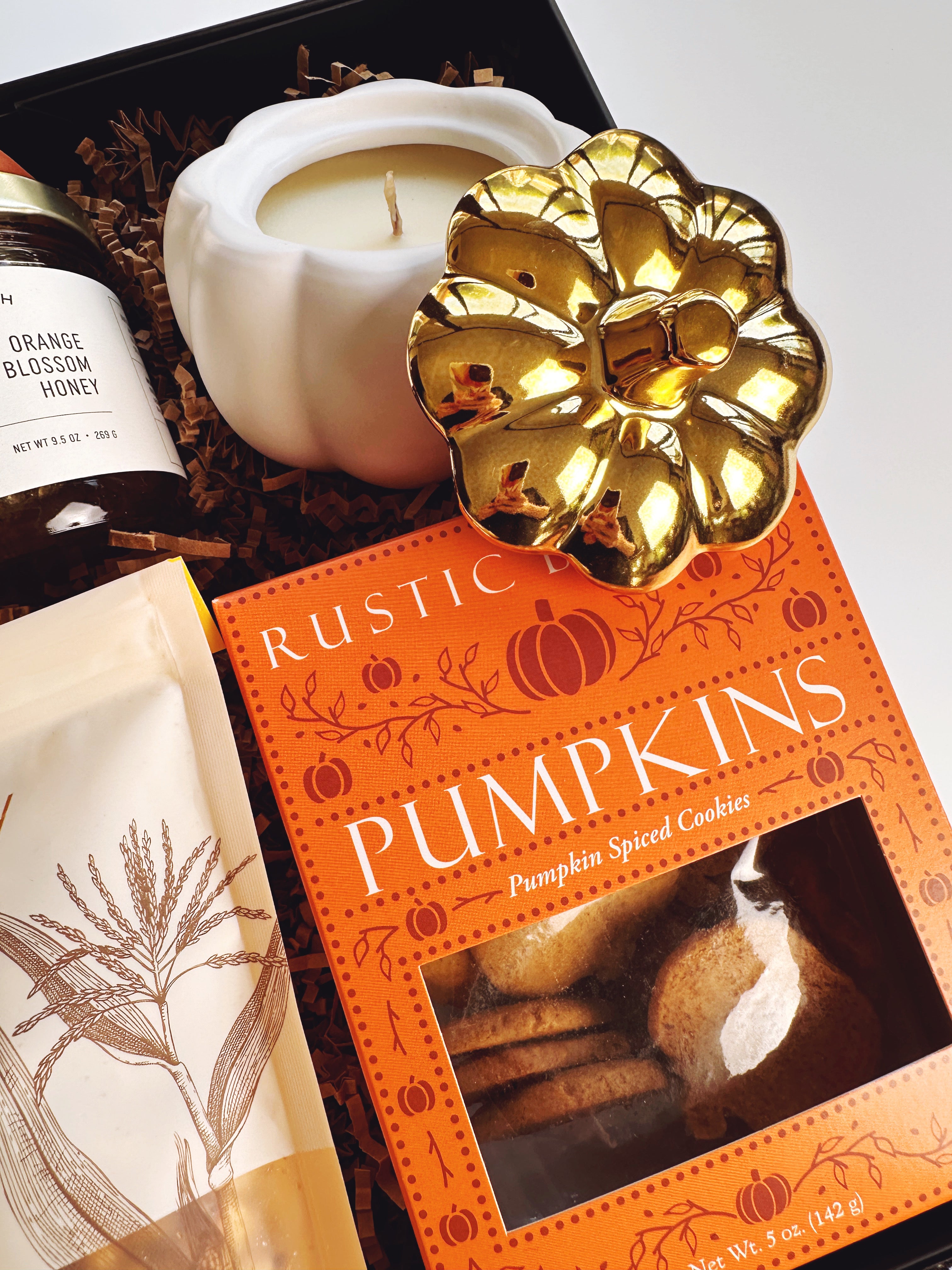 A gift box arrangement featuring a box of Rustic Bakery Pumpkin Spice Cookies, a white pumpkin-shaped candle,  and a jar of orange blossom honey.