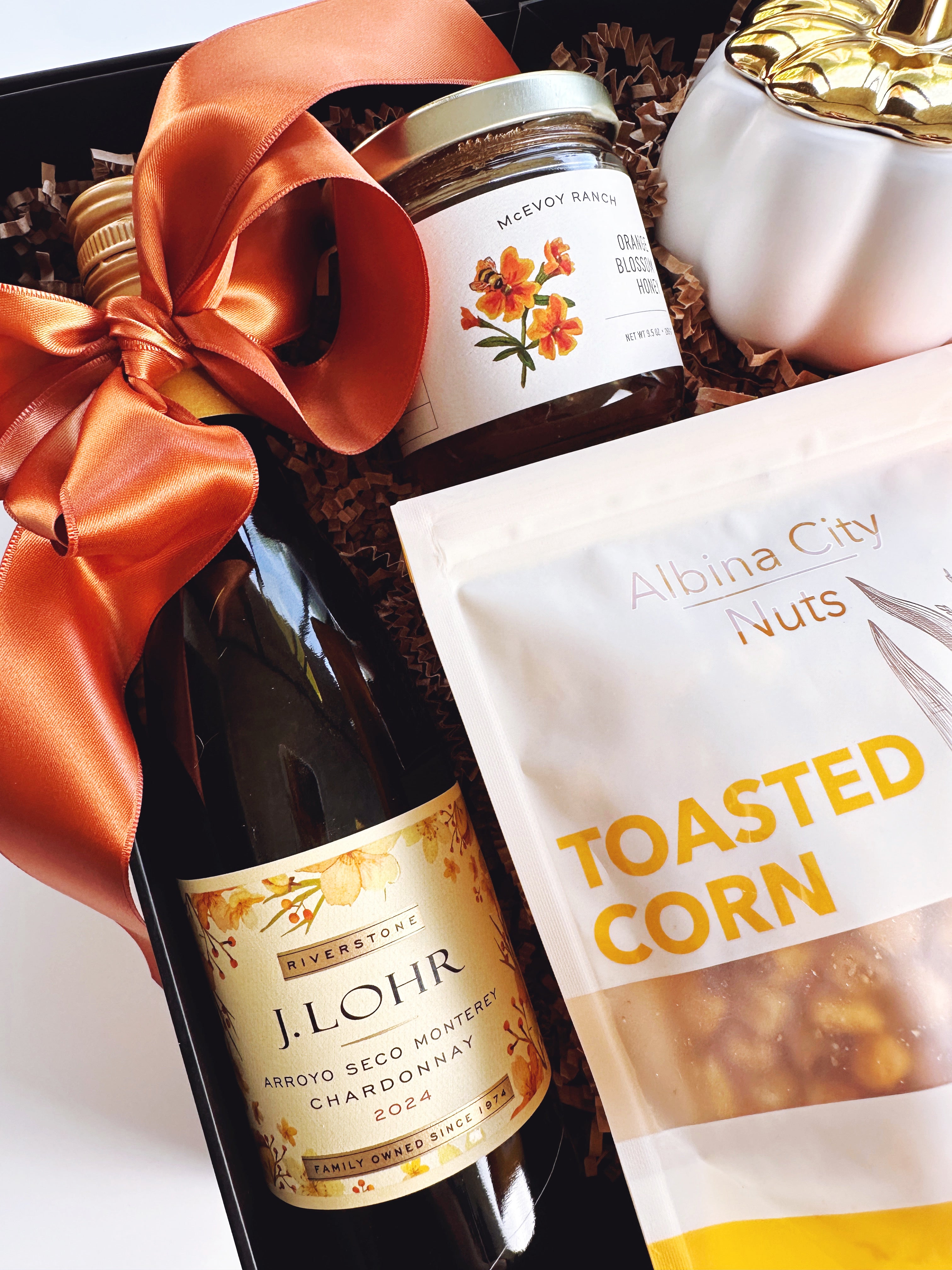 A curated gift basket or hamper containing a bottle of J. Lohr iChardonnay 2024, a jar of honey, a bag of Albina City Nuts Toasted Corn, and a white decorative pumpkin candle all arranged in a black box with a large rust colored satin ribbon.