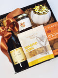 An autumn-themed gift basket set from with Albina Roasted Corn, featuring a bottle of chardonnay, a jar of honey, pumpkin cookies, a small white pumpkin Candle, and a Rust Colored ribbon, all arranged in a black box with shredded paper around the items.