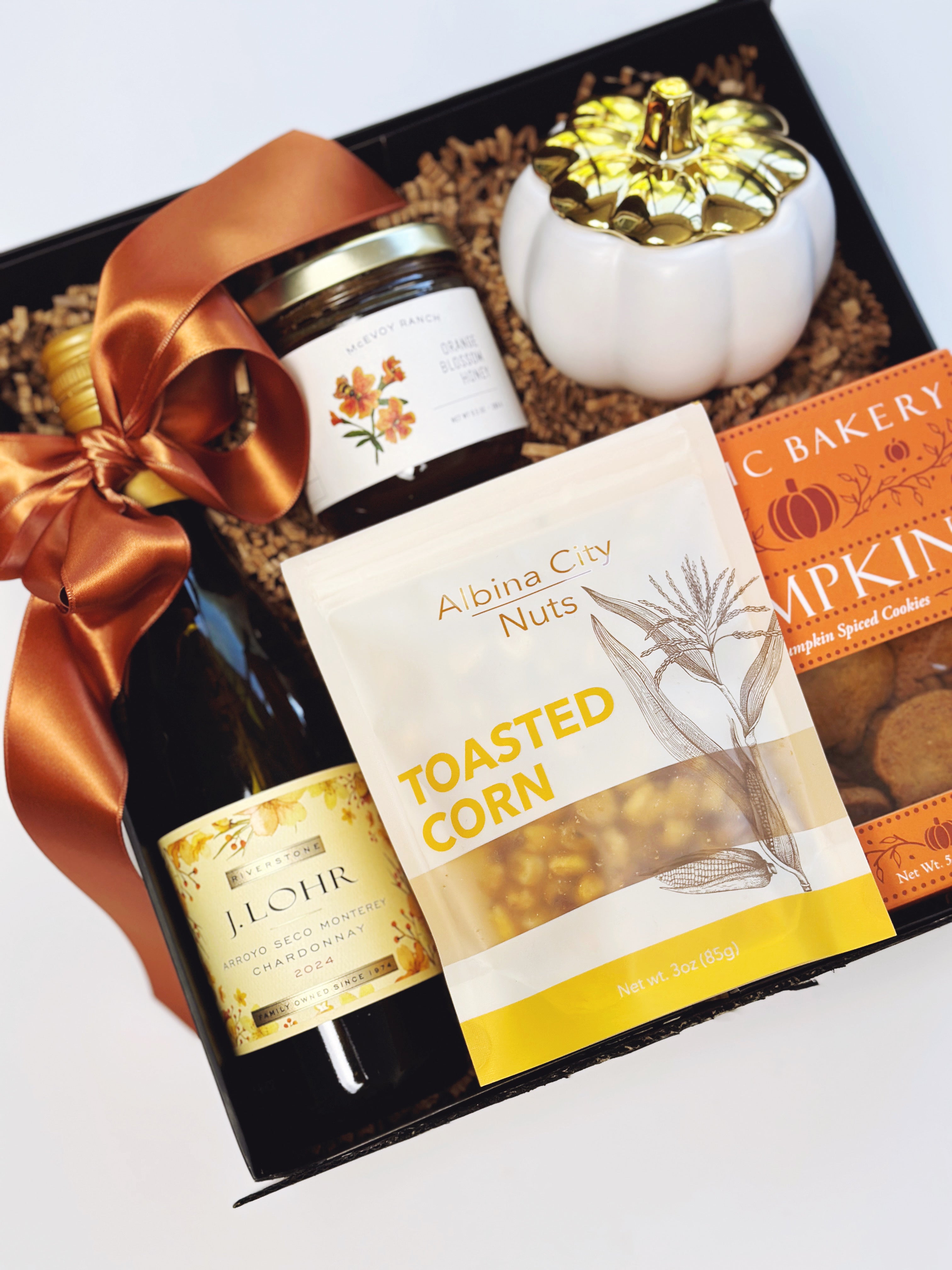 An autumn-themed gift basket set from with Albina Roasted Corn, featuring a bottle of chardonnay, a jar of honey, pumpkin cookies, a small white pumpkin Candle, and a Rust Colored ribbon, all arranged in a black box with shredded paper around the items.