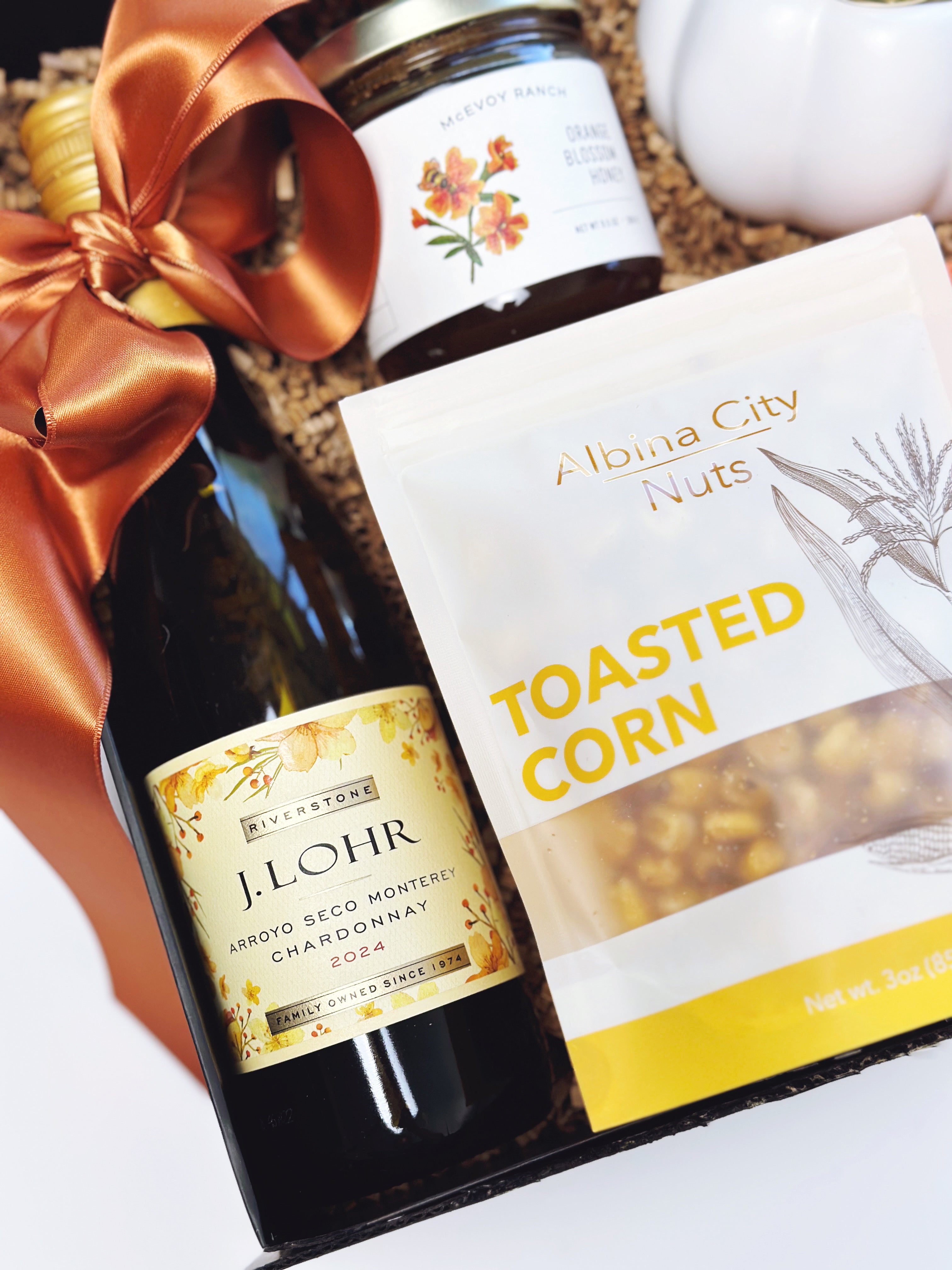 A gift basket arrangement featuring a bottle of J. Lohr Arroyo Seco Monterey Chardonnay 2024, a bag of A Ibina City Nuts Toasted Corn, a jar of McEvoy Ranch Honey, and a large copper-orange ribbon bow, set in a basket with shredded paper filler.