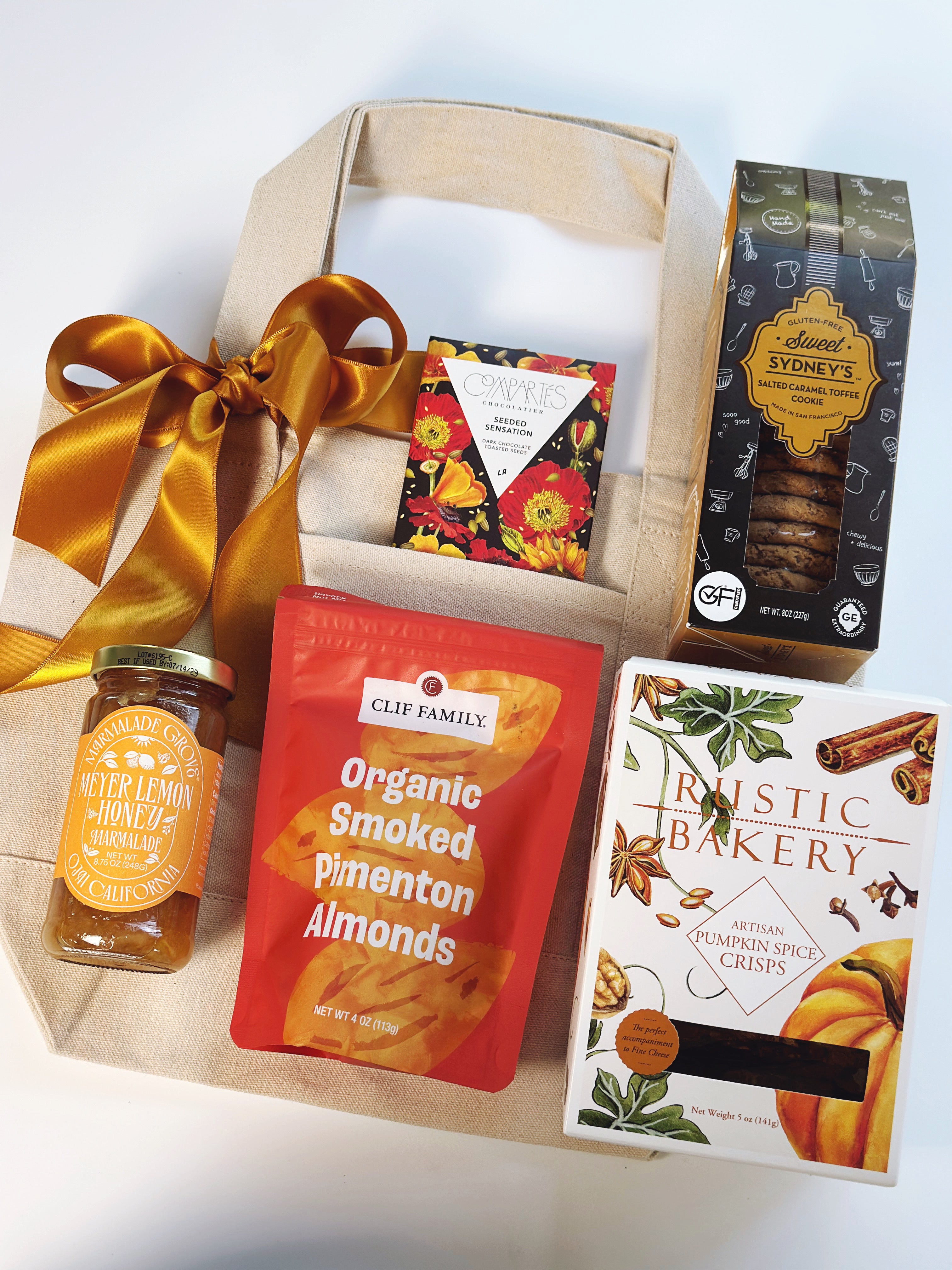 A curated gift basket set on a white background, featuring a beige canvas tote bag with a large gold ribbon, a jar of Clif Family Pimento Honey, a red bag of Clif Family Organic Smoked Pimento Almonds, a box of Rustic Bakery Pumpkin Spice Crispins, and a box of Australian Sydney Bakes chocolate chip cookies.