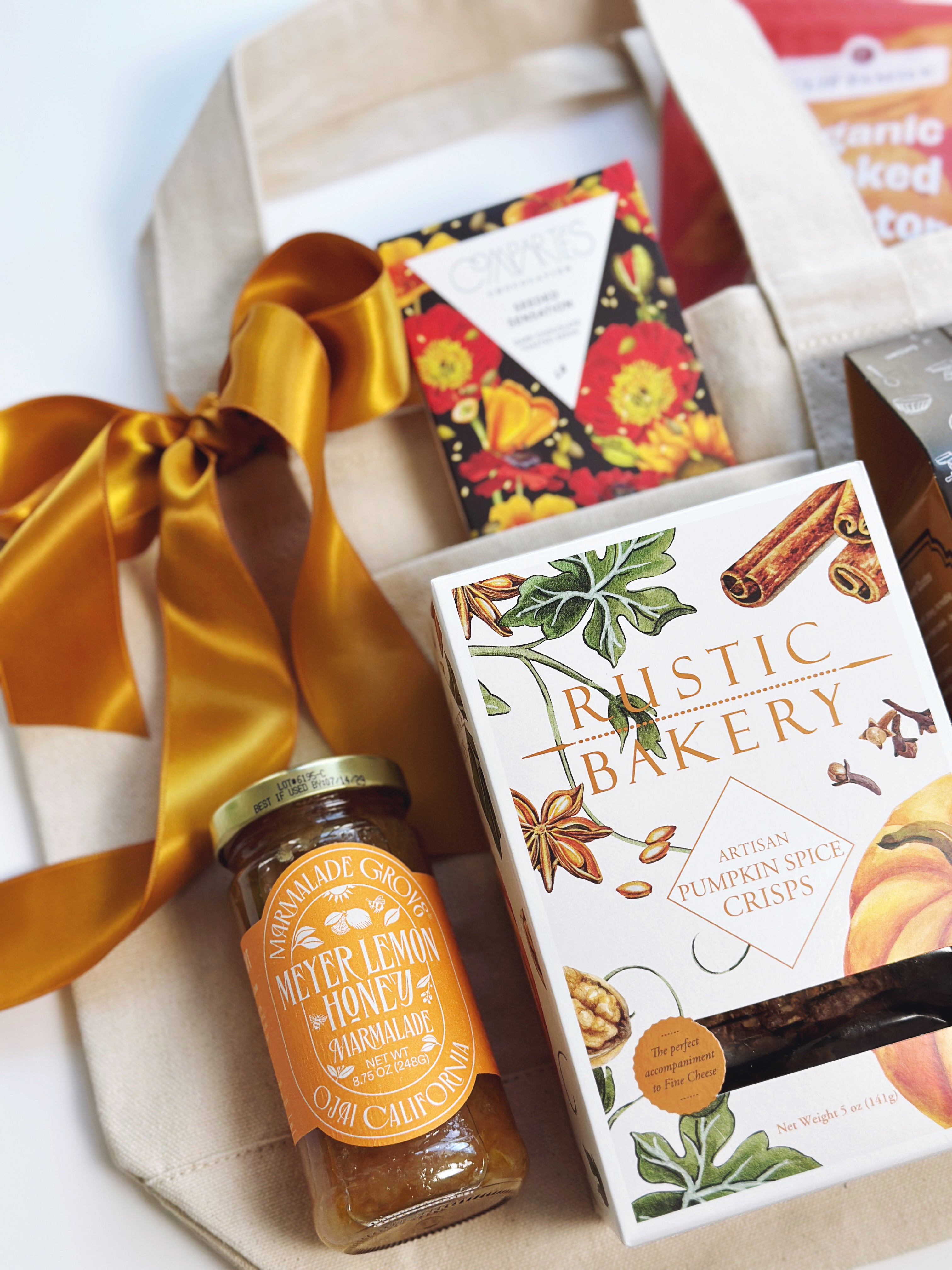 A canvas gift bag arrangement featuring a jar of Meyer Lemon Honey jam, a box of Rustic Bakery artisan pumpkin spice crisp, a floral-patterned box, and a beige tote bag tied with a gold ribbon on a white background.