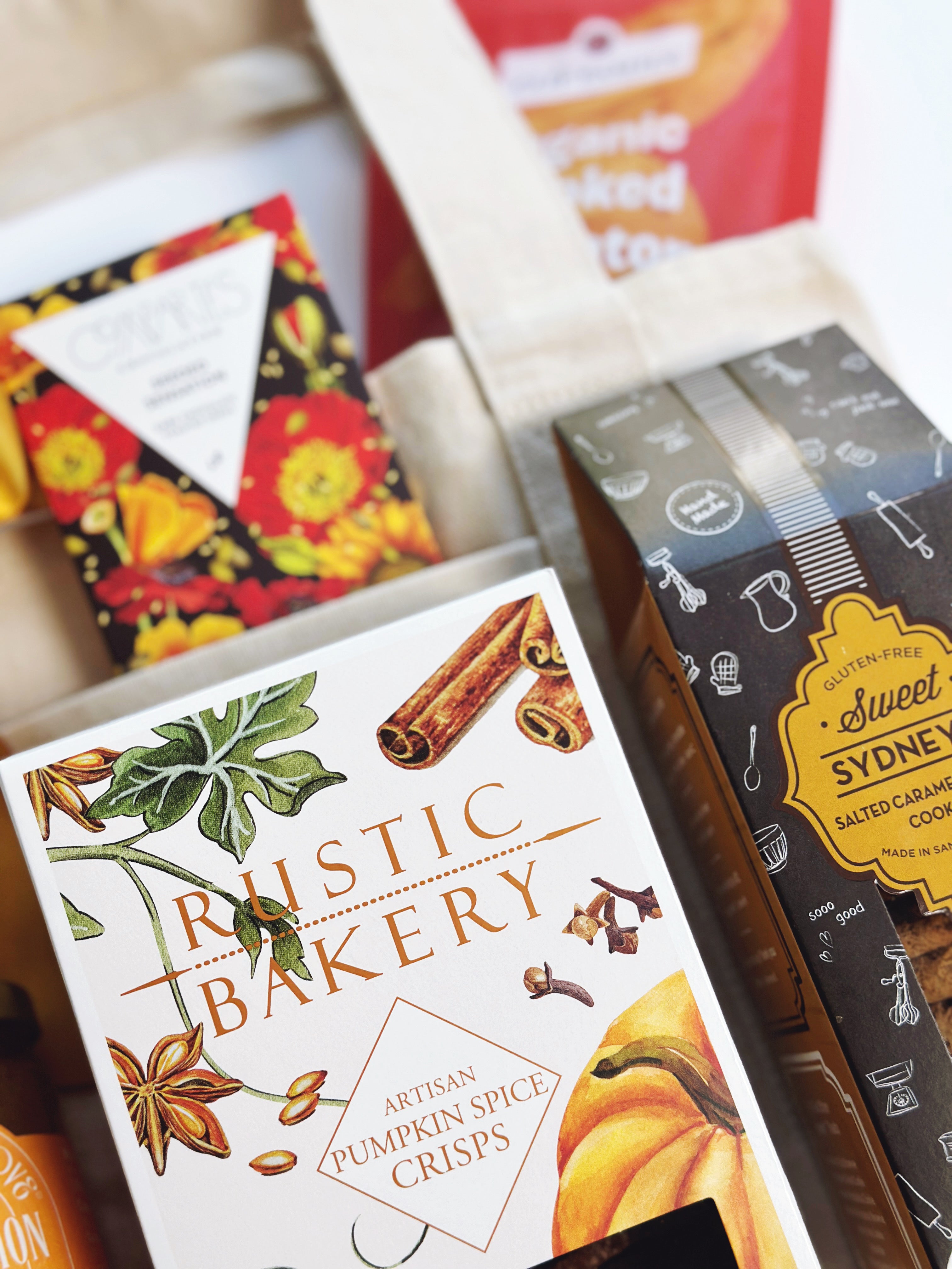 A close-up of packaged baked goods in a gift-style arrangement. The most prominent item is a box of Rustic Bakery Artisan Pumpkin Spice Crisps with botanical illustrations and a pumpkin graphic.