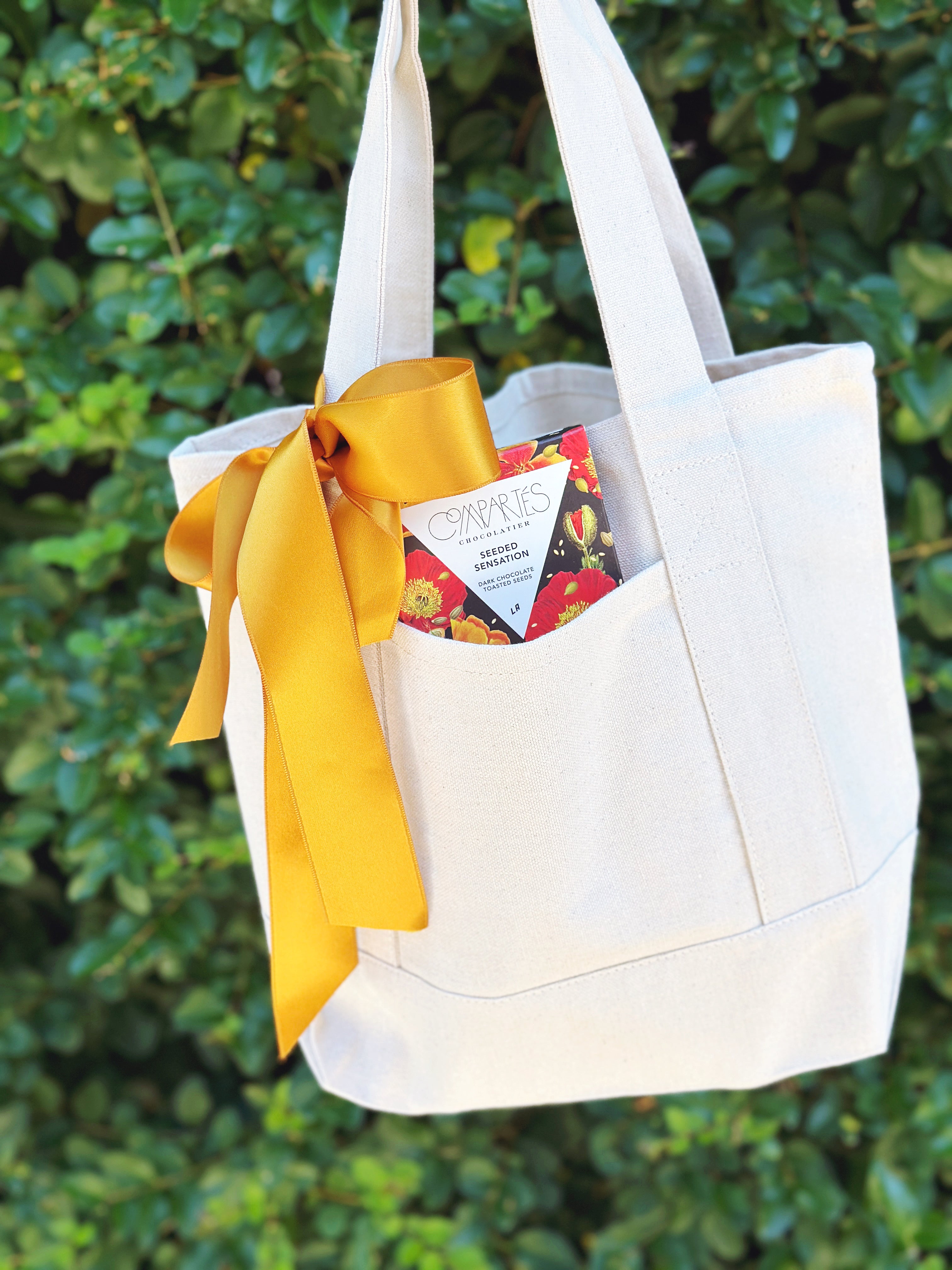An off-white canvas tote bag with a large yellow satin ribbon bow, shown hanging outdoors against a background of green foliage. A chocolate bar is tucked into one of the front pockets.