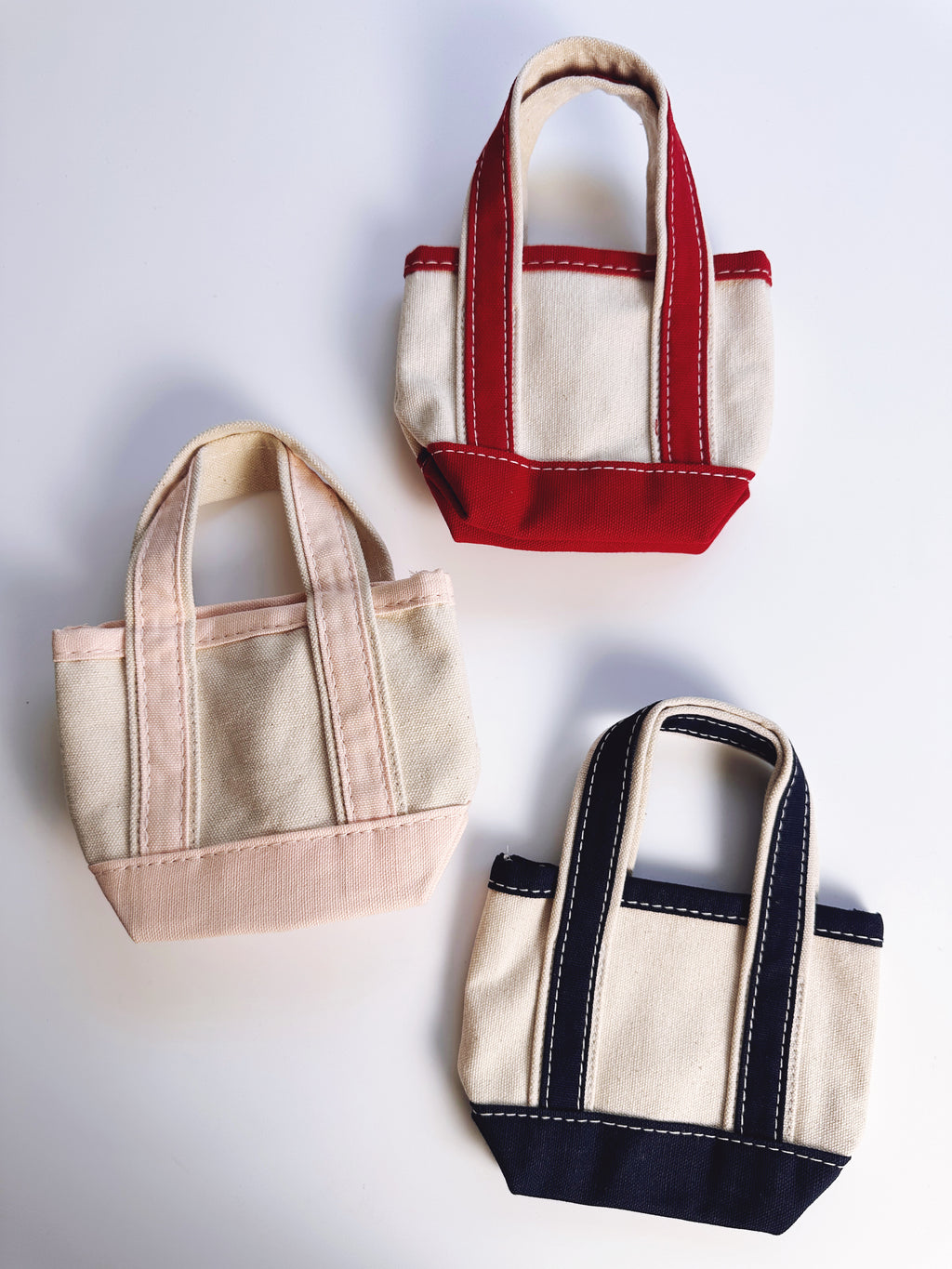 Three small canvas tote bags arranged on a light gray background, each with cream fabric and contrasting trim in red, blush pink, and navy blue.