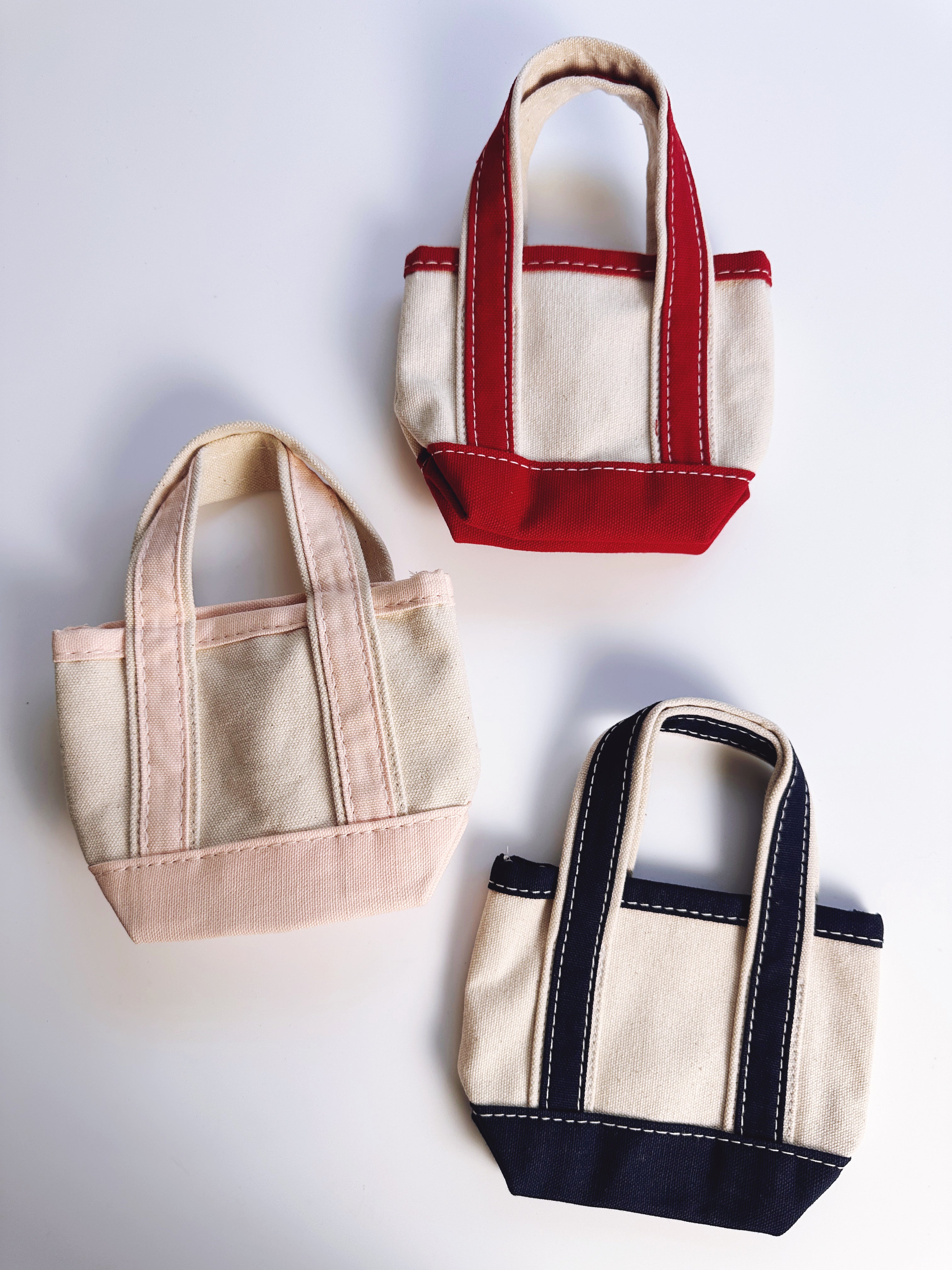 Three small canvas tote bags arranged on a light gray background, each with cream fabric and contrasting trim in red, blush pink, and navy blue.