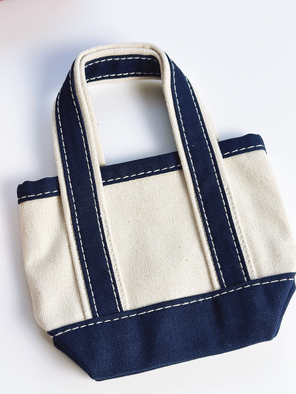A small canvas tote bag with a cream-colored body and navy blue trim, featuring white contrast stitching and two top handles, shown on a plain white background.