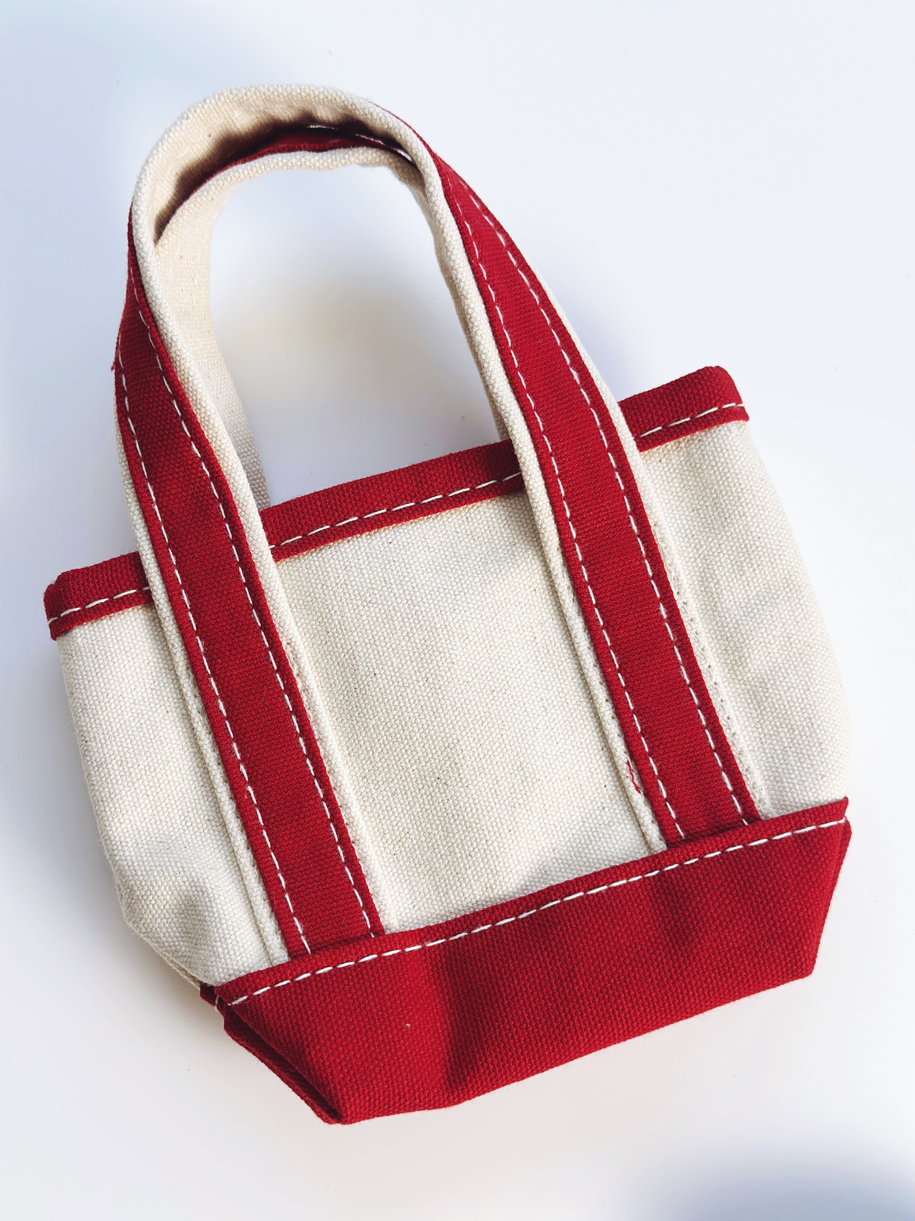 A small canvas tote bag with cream-colored panels and red trim, featuring double shoulder straps and white contrast stitching, photographed on a clean white background.