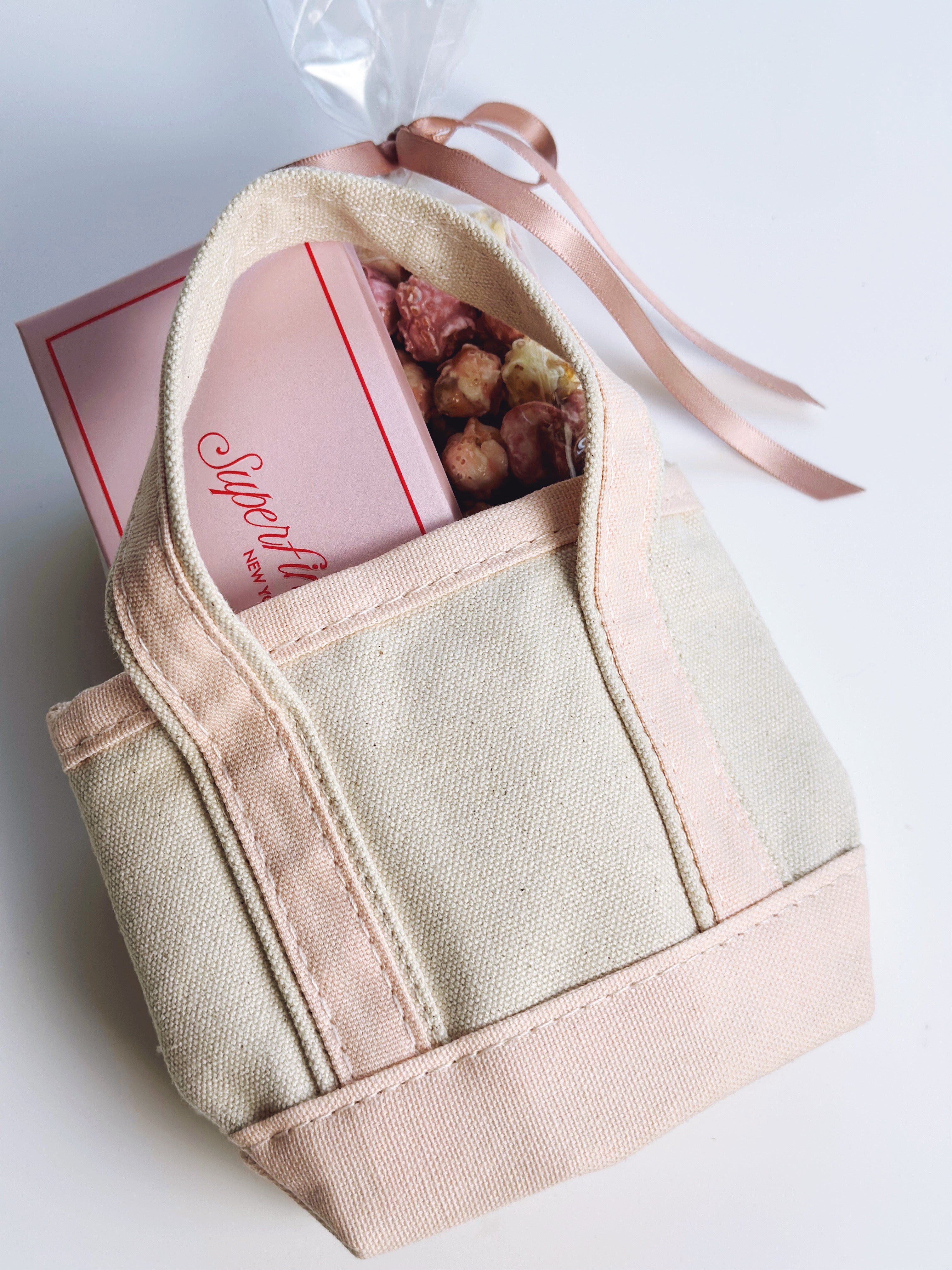 A small canvas tote bag in warm beige and blush pink, shown open with a pink chocolate bar and popcorn inside, tied with a matching pink ribbon on a white background.