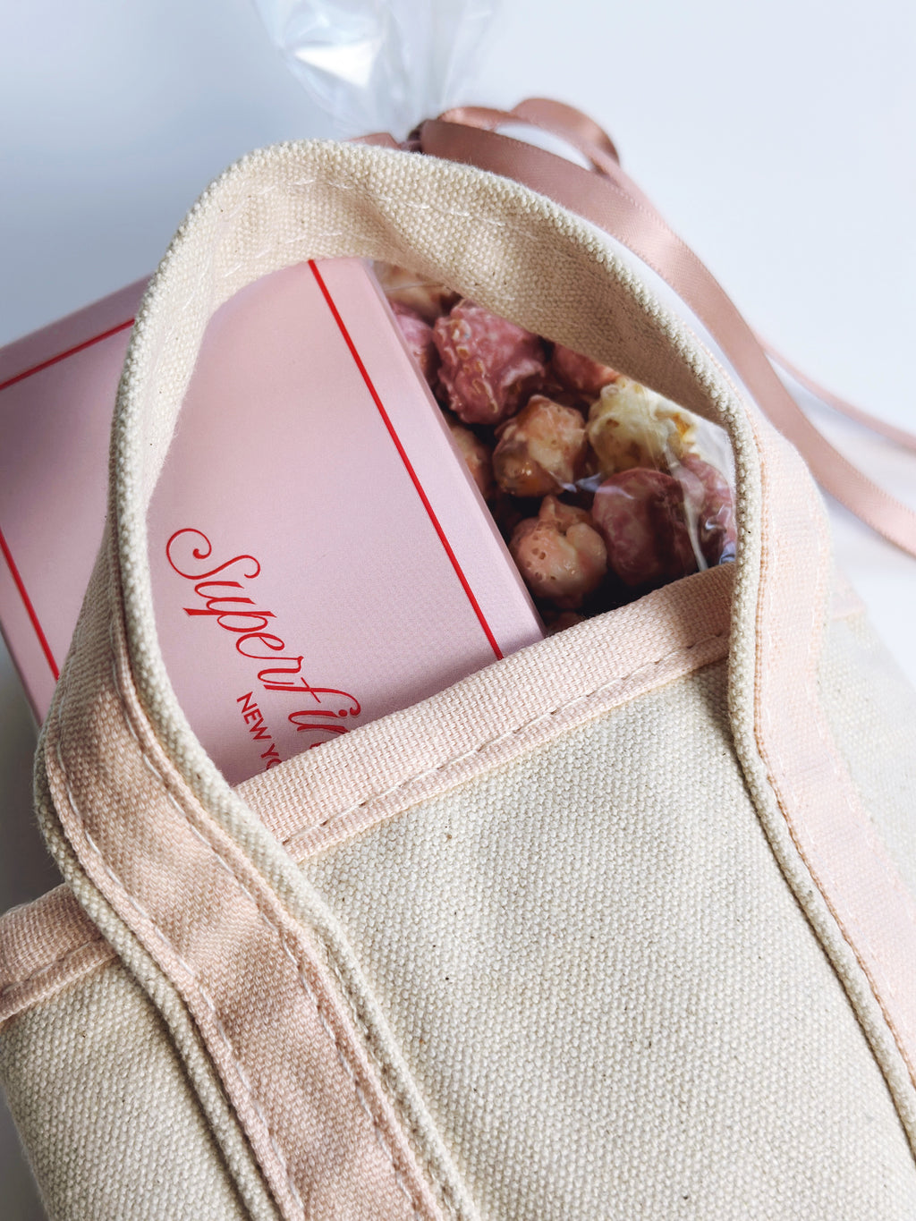 A close-up of a beige canvas tote bag with a pink ribbon, showing a pink chooclate bar and popcorn in a slim bag tucked inside.