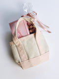 A small canvas tote bag in natural beige and blush pink, shown from a top-down angle on a white background. A pink gift box with a pink ribbon is tucked inside the bag.