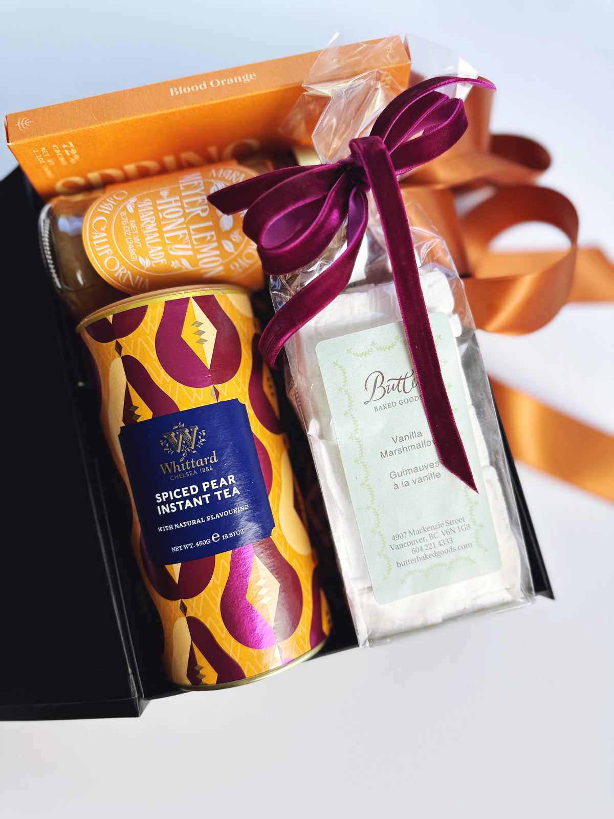 A gift box containing a can of Whittard Spiced Pear Instant Tea, a jar of marmalade, a packaged vanilla marshmallows, and an orange chocolate bar,  tied together with a dark purple velvet ribbon.