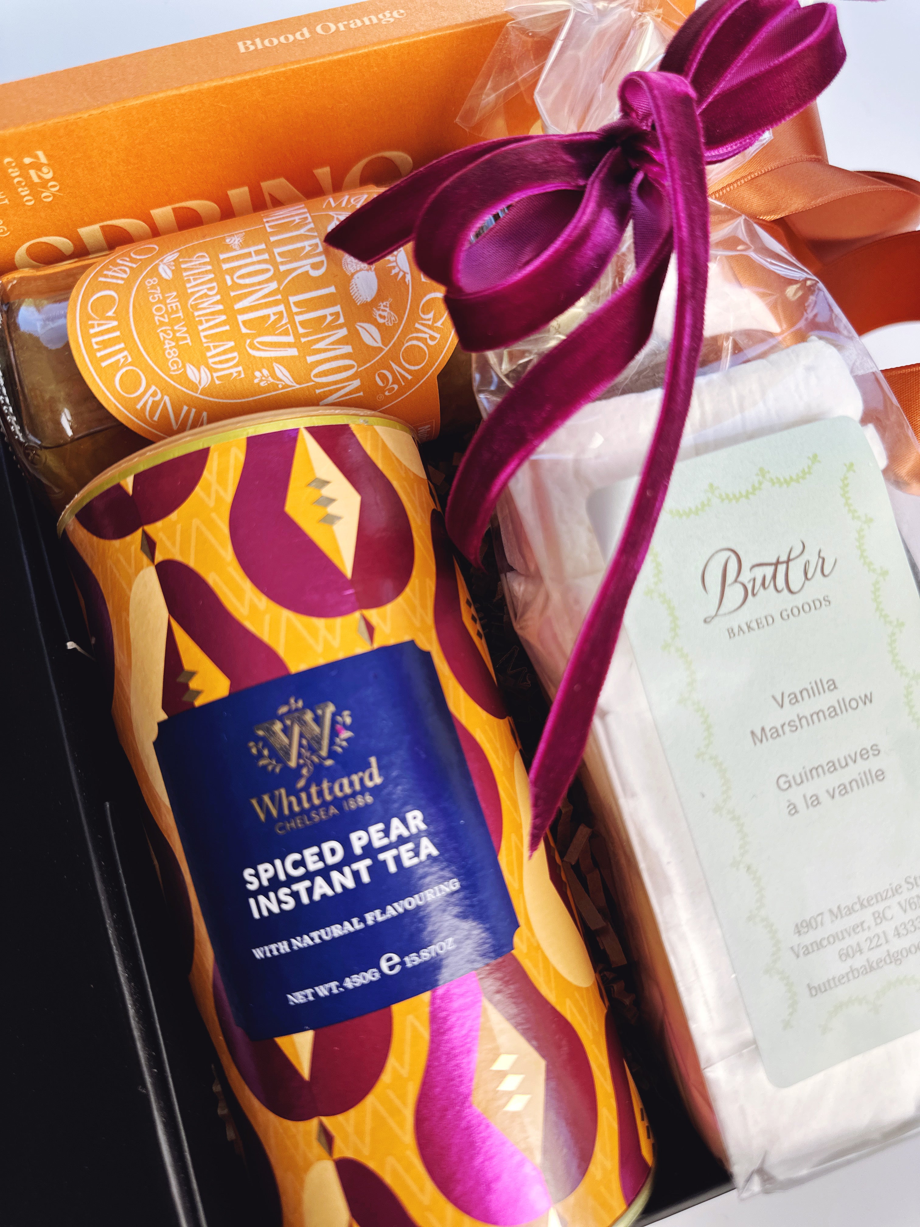 A gift box containing a can of Whittard Spiced Pear Instant Tea, a package of Butter Vanilla Marshmallow, and a bright magenta velvet ribbon bow, set against an orange background.