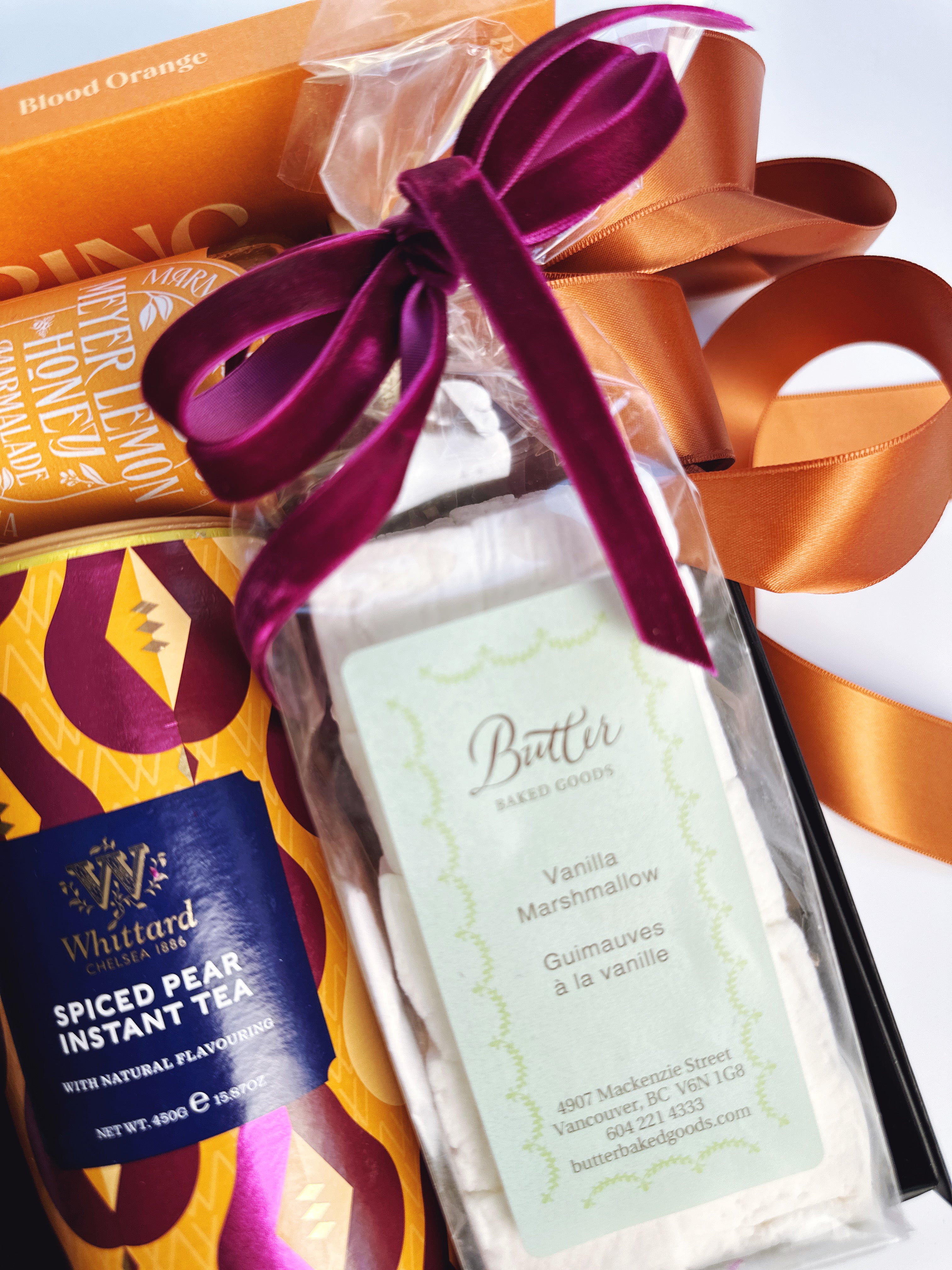 A gift basket arrangement featuring a bag of Butter Baked Goods Vanilla Marshmallow butter tied with a purple ribbon, alongside a box of Whittard Spiced Bear Instant Tea and an orange box, all set against a clean white background.