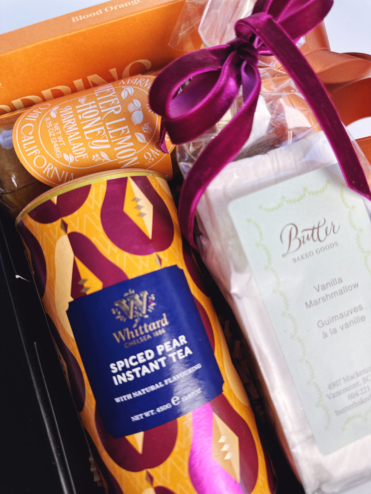 A gift basket or hamper containing a can of Whittard Spiced Pear Instant Tea, a package of Butter Baked Goods Vanilla Marshmallow butter, and a wrapped item tied with a purple ribbon, all arranged in a black box.