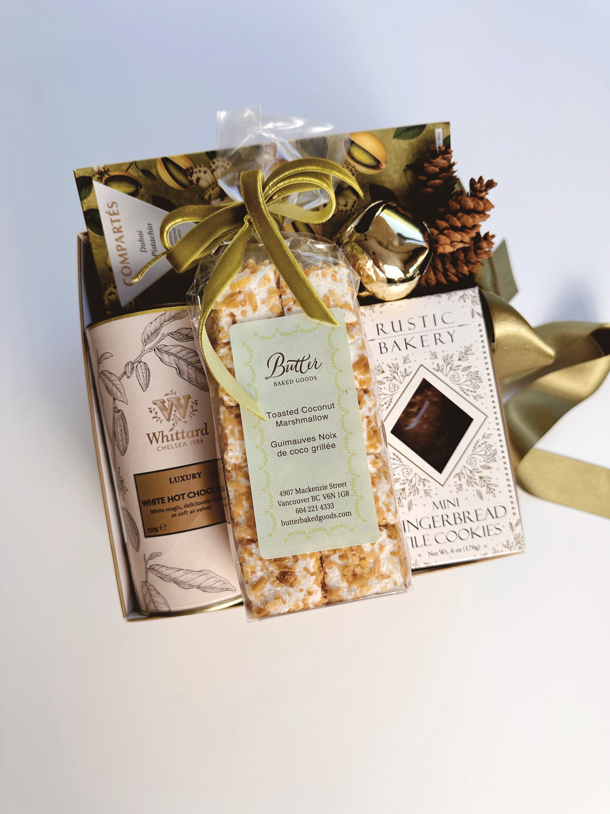 A gourmet gift box featuring a Whittard White Hot Chocolate, a bag of Butter  Toasted Coconut Marshmallows and a box of Rustic Bakery Mini Gingerbread Cookies, tied together with a green ribbon and decorated with pinecones on a light background.