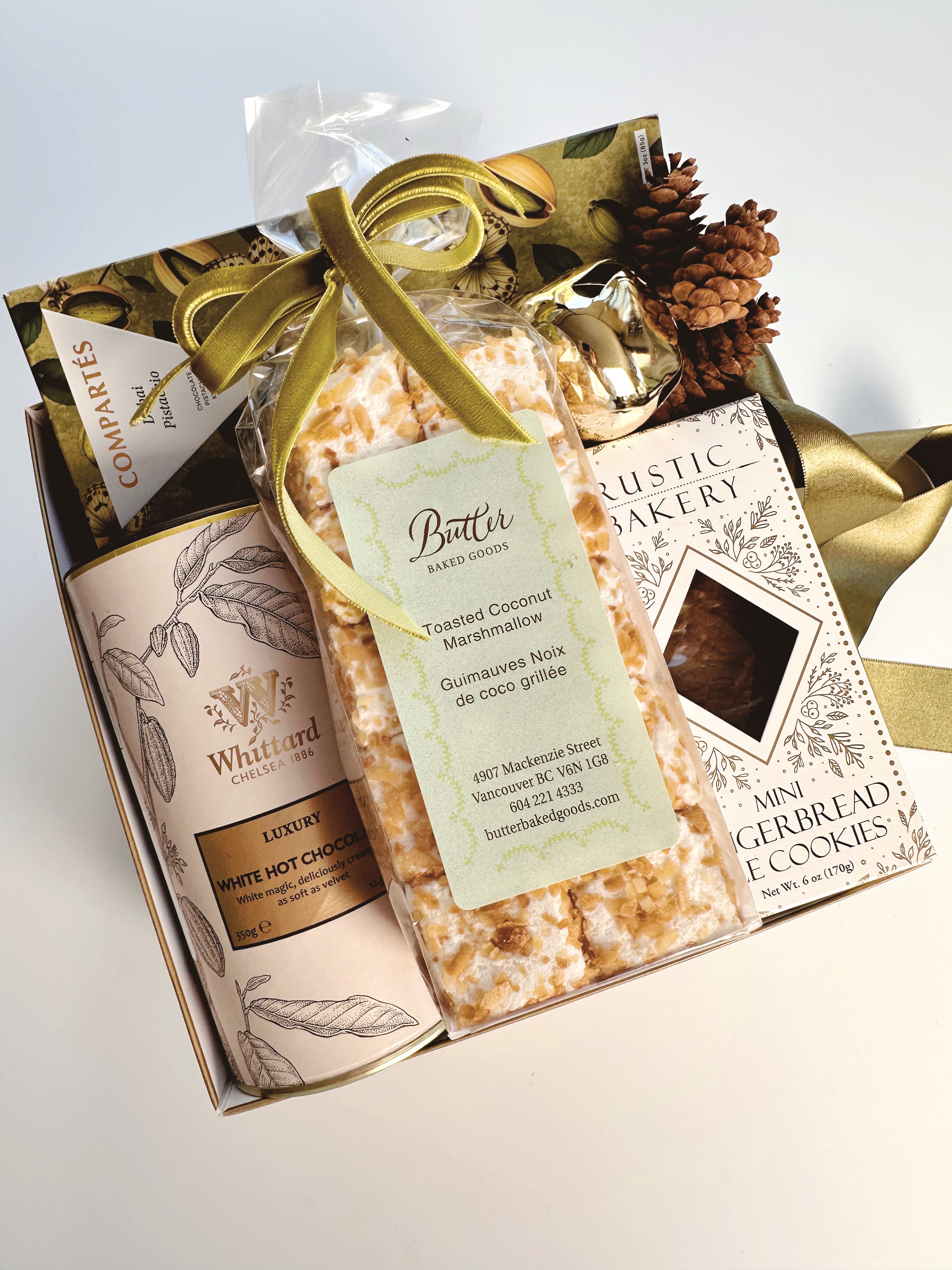 A gourmet gift basket with Whittard white chocolate cocoa and Rustic Bakery gingerbread, featuring a bag of toasted coconut marshmallows tied with a green velvet ribbon.  Tucked at the top is a Dubai Pistachio Chocolate Bar.