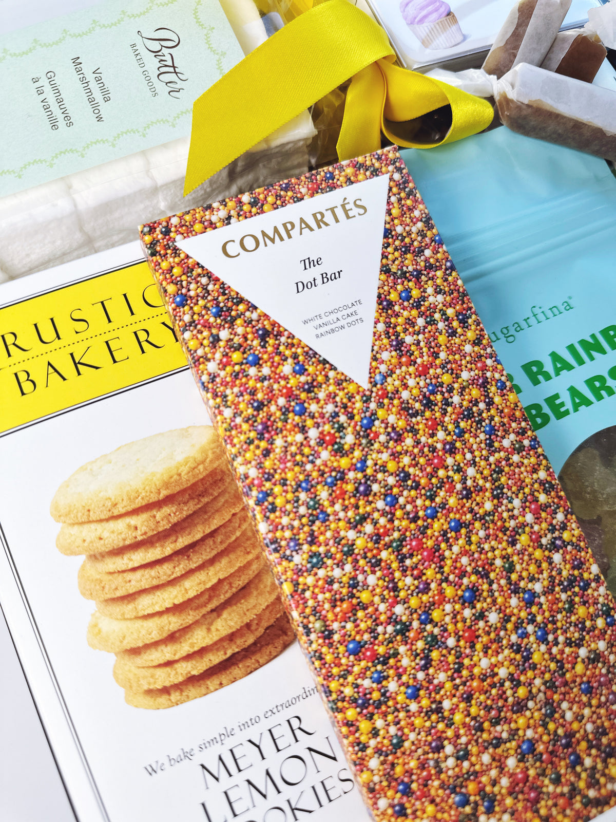 A close-up of a Compartés 'The Dot Bar' chocolate box covered in multicolored sprinkles, surrounded by other gift items including a Rustic Bakery Meyer Lemon Cookies.