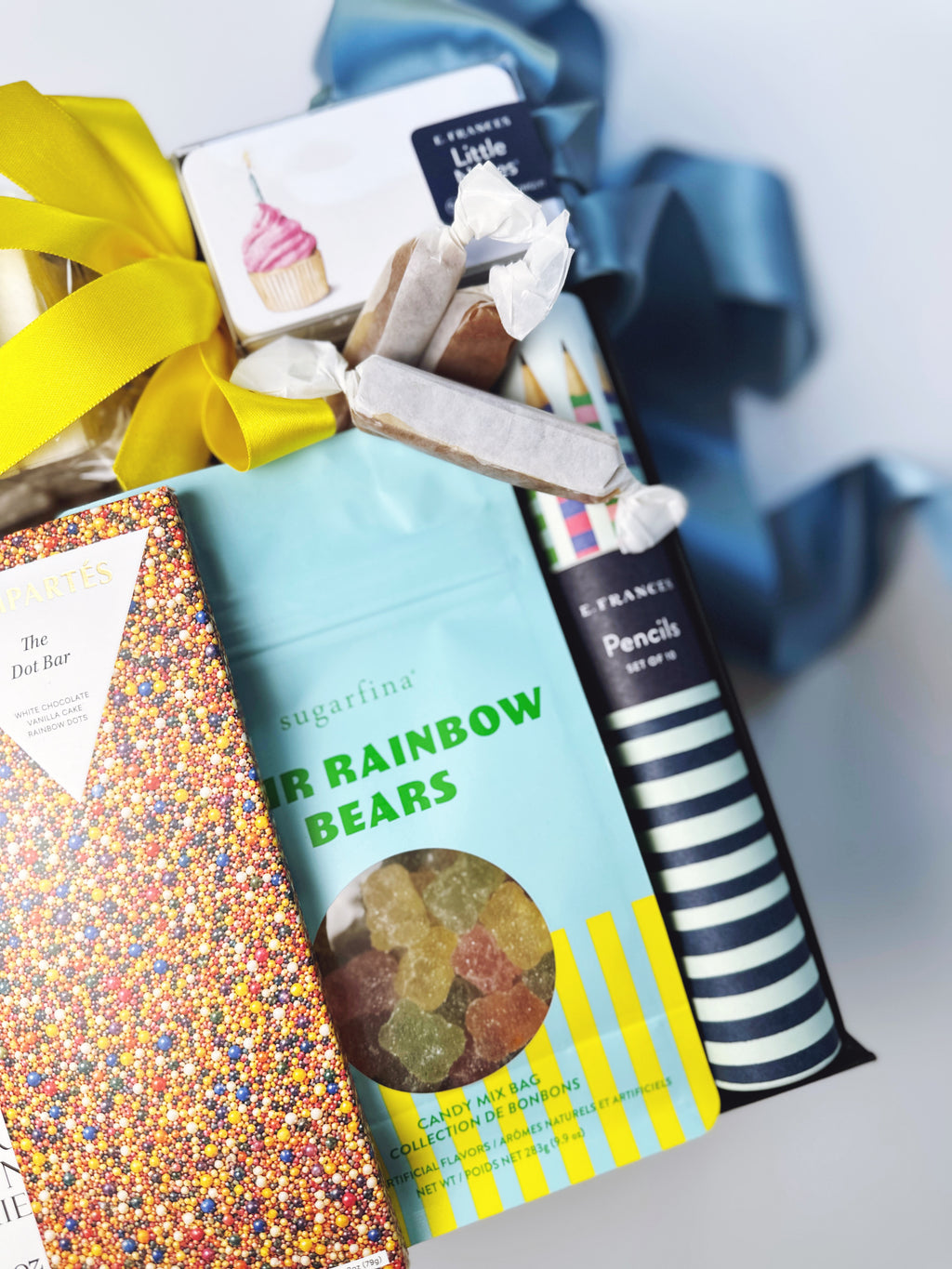 A gift basket filled with assorted snacks and stationery, including a bag of sugarfina Rainbow Bears gummy bears, a striped pencil box, a cupcake-themed card, and a yellow ribbon bow, photographed on a light gray background.
