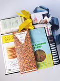 A curated gift basket filled with assorted snacks and treats, including a box of Rustic Bakeries Meyer Lemon Cookies, a Compartés chocolate bar, a bag of Rainbow Bears gummy bears, and a pencil, tied with a yellow ribbon and blue ribbon.