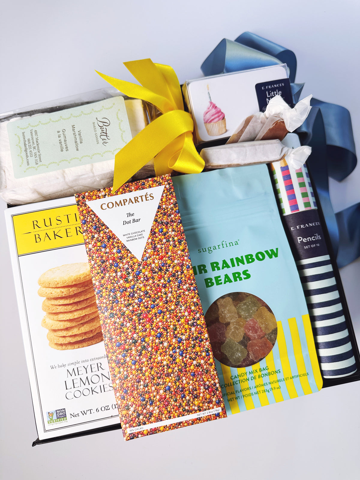 A curated gift basket filled with assorted snacks and treats, including a box of Rustic Bakeries Meyer Lemon Cookies, a Compartés chocolate bar, a bag of Rainbow Bears gummy bears, and a pencil, tied with a yellow ribbon and blue ribbon.
