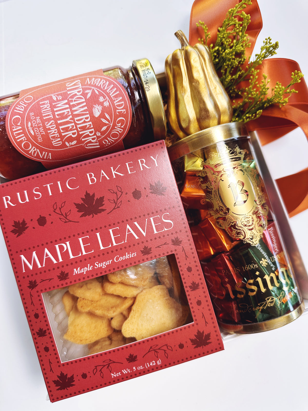 A styled gift assortment featuring a red box of Rustic Bakery Maple Leaves maple sugar cookies, a jar of strawberry marmalade, and Bissinger's Chocolate Caramels.