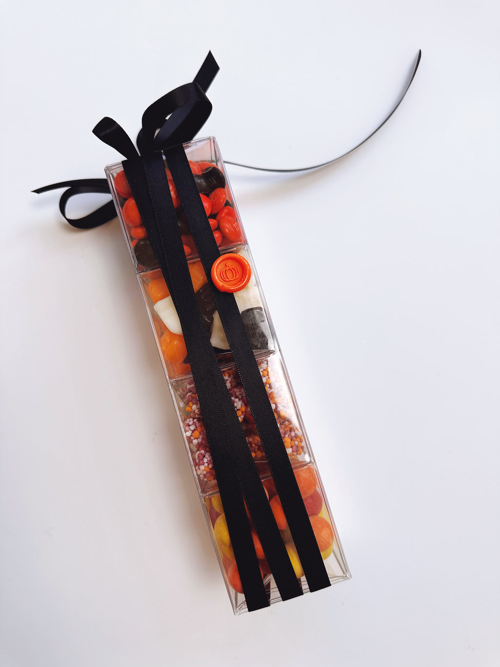 A clear rectangular candy box filled with assorted Halloween-themed candies in orange, black, and white, tied with a black ribbon and bow on a plain white background.