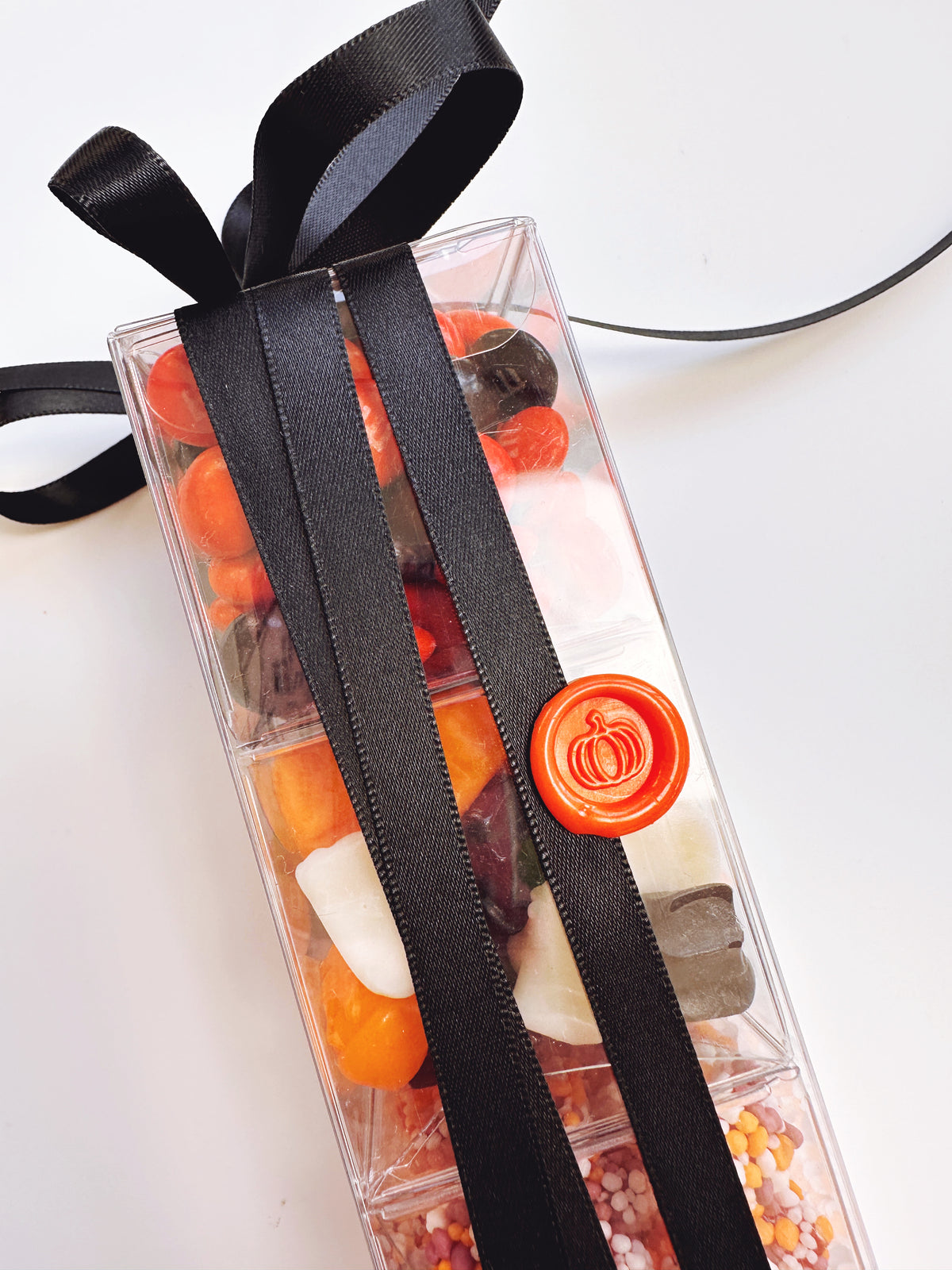 A clear rectangular gift box filled with assorted colorful candies and sweets, tied with a wide black ribbon and sealed with a round orange wax stamp, photographed on a white background.