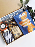 An open kraft gift box arranged with multiple food items and a striped jar of McEvoy Ranch olive oil on a white surface. Visible products include a blue Clif Family Organic Tamari Glazed Mixed Nuts bag, a jar of Paloma de Colores popcorn kernels, a bottle of Souse Salt, and a wrapped Salami.