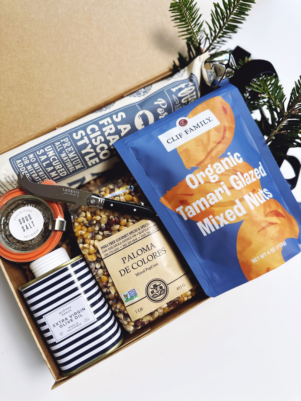 An open kraft gift box arranged with multiple food items and a striped jar of McEvoy Ranch olive oil on a white surface. Visible products include a blue Clif Family Organic Tamari Glazed Mixed Nuts bag, a jar of Paloma de Colores popcorn kernels, a bottle of Souse Salt, and a wrapped Salami.