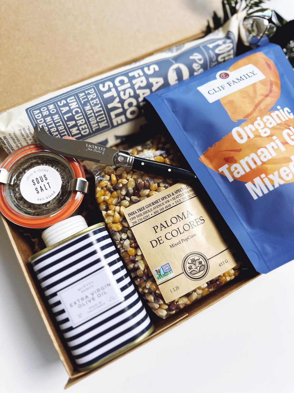 An open gift box filled with assorted packaged snacks and food items, including a blue bag of Clif Family Organic Tamari Cereal Mix, a striped jar of Olive Oil and a bag of Paloma de Colores  popcorn, arranged on a light surface.