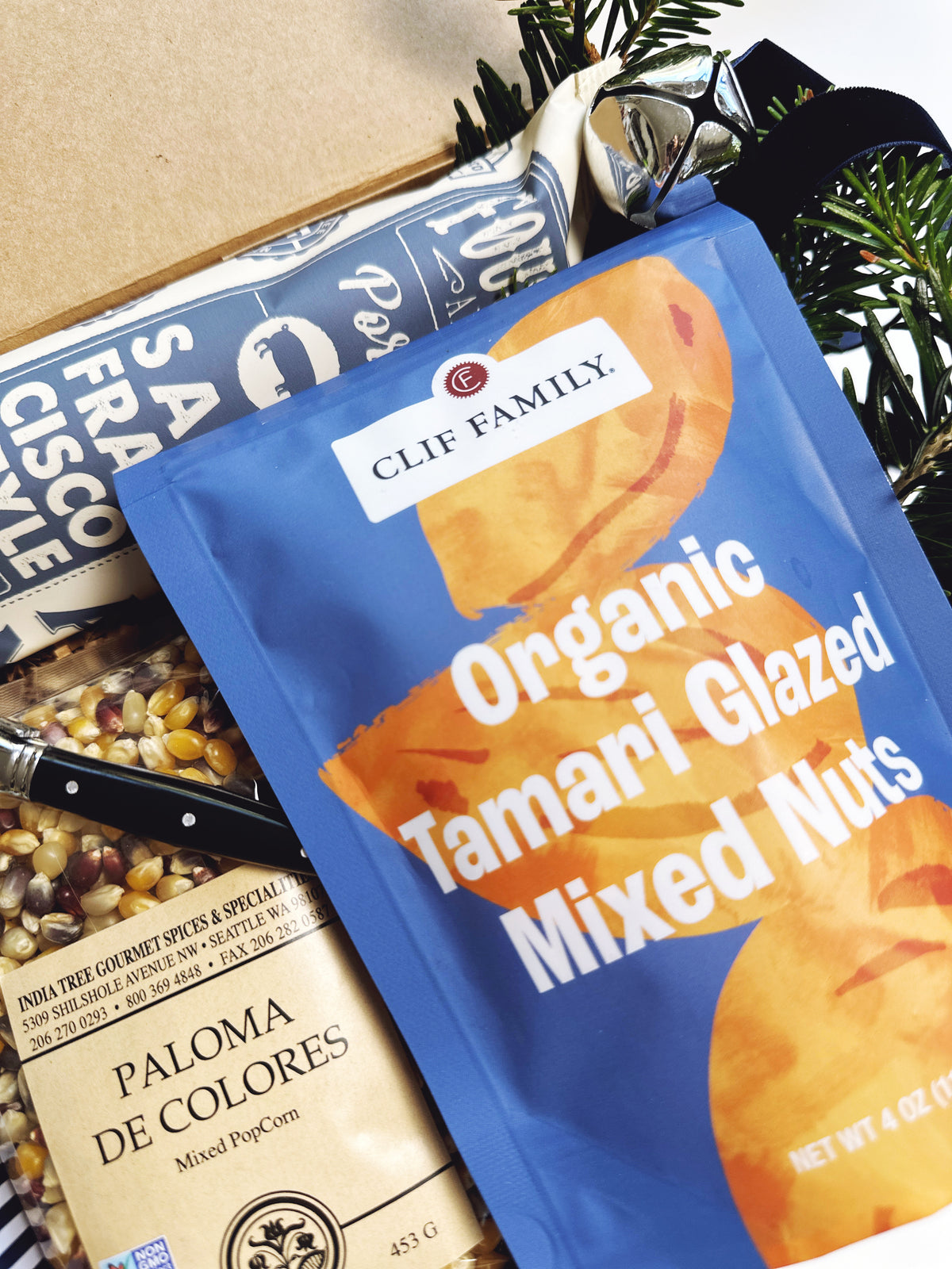 A Clif Family Organic Tamari Glazed Mixed Nuts bag in blue packaging, styled inside a gift box with other gourmet snacks and a decorative black ribbon.