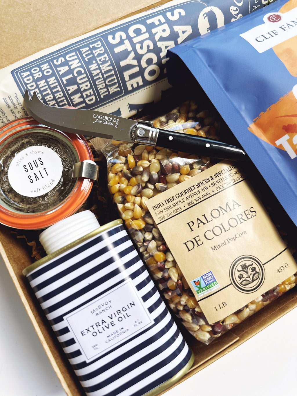 A curated gourmet gift box filled with various food items, including a can of McEvoy Ranch Extra Virgin Olive Oil, a jar of Clif Family Nuts, Sous Salt, a bag of Paloma de Colores popcorn.