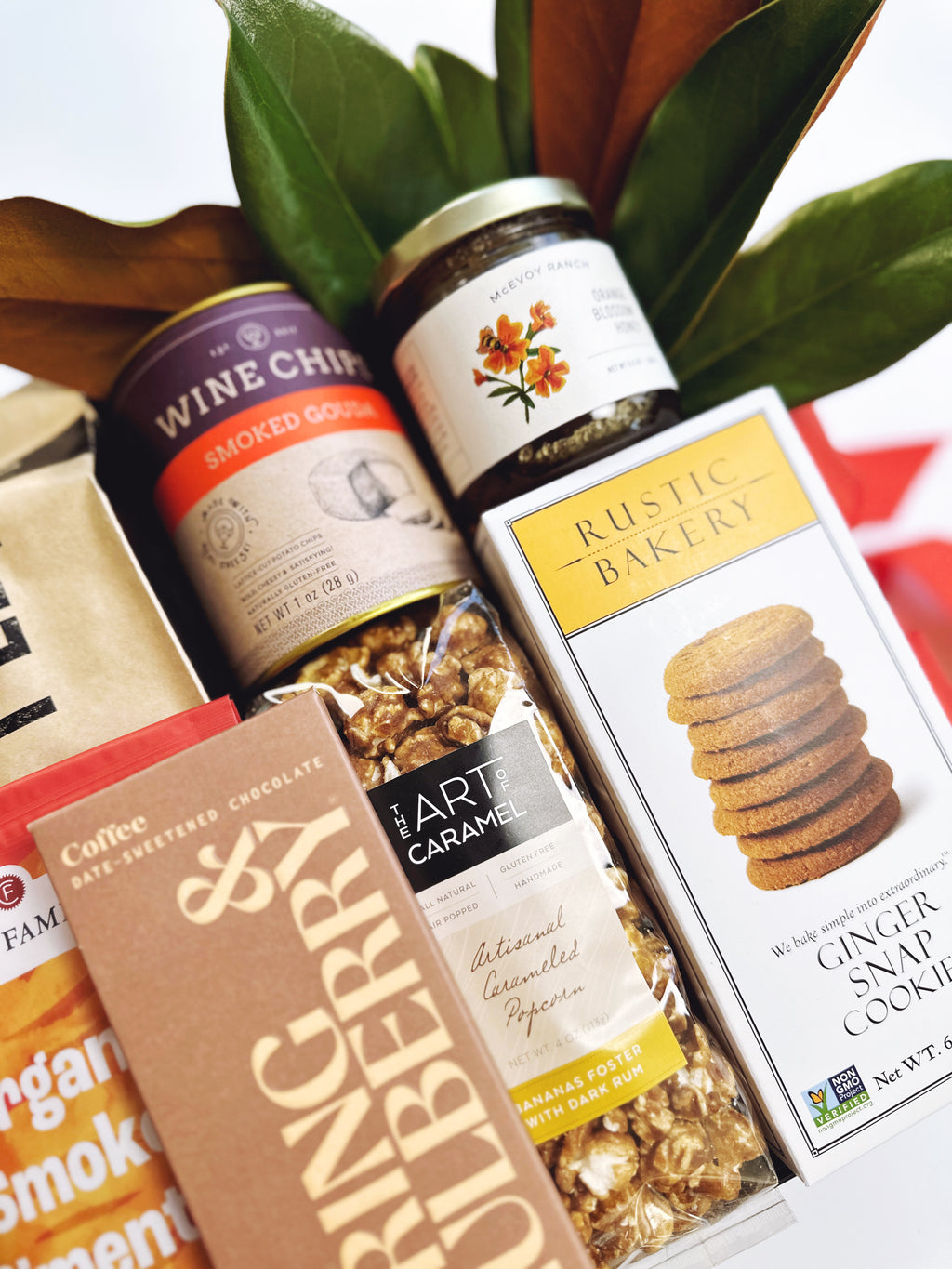 A curated gift basket or box filled with assorted packaged snacks and baked goods, including a jar of wine chip seasoning, a box of Rustic Bakery ginger snap cookies,  Caramel Corn, Wine Chips and Snacks
