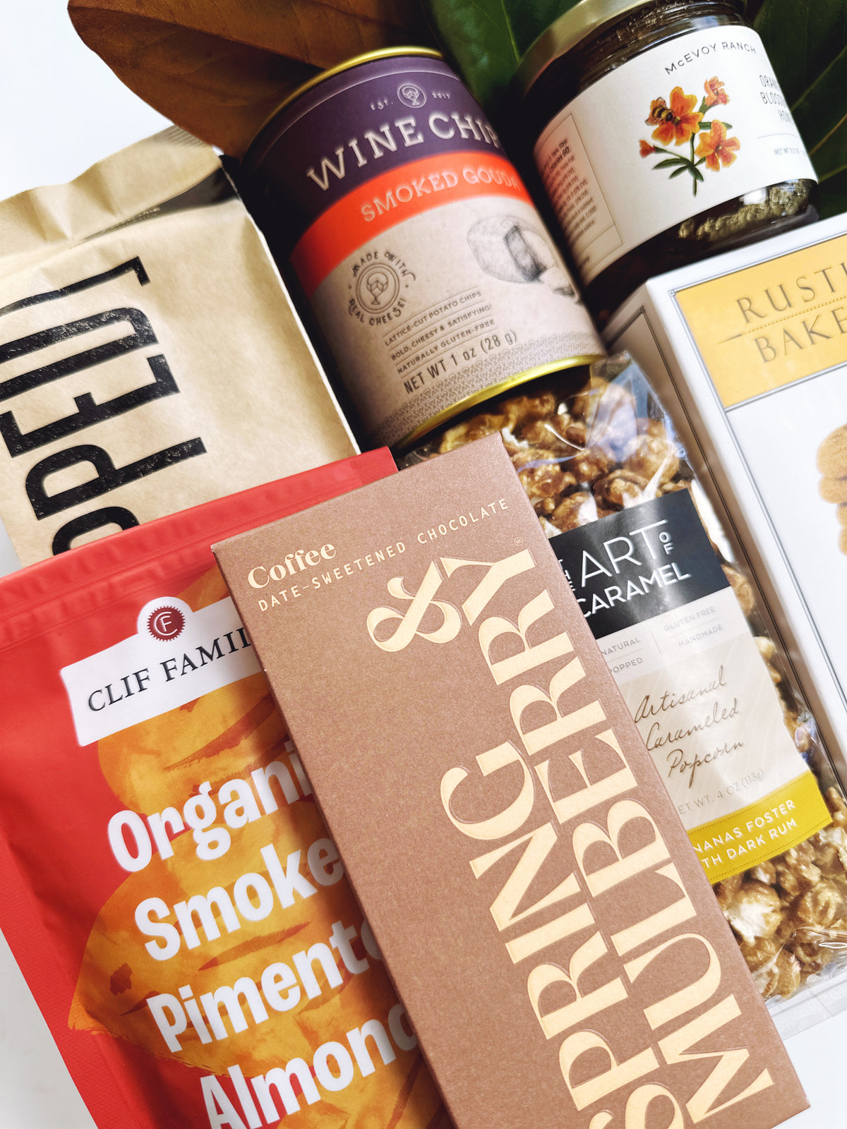 A curated gift basket or snack assortment featuring a bag of Clif Family Organic Smokeless Pimento Almonds, a brown paperboard box of Nutriberry & Gerbera coffee dried sweetened chocolate, a can of Wine Chips Smoked Gourmet, a jar of Marzipan, and a bag of Rustic Baked Granola.